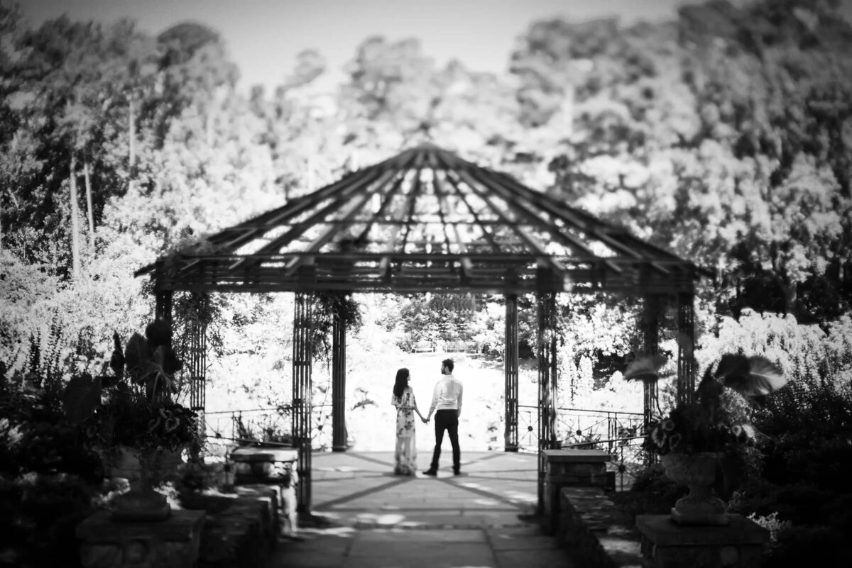 Black and white image of a couple standing in a pergola, with their silhouettes framed by the structure's intricate patterns