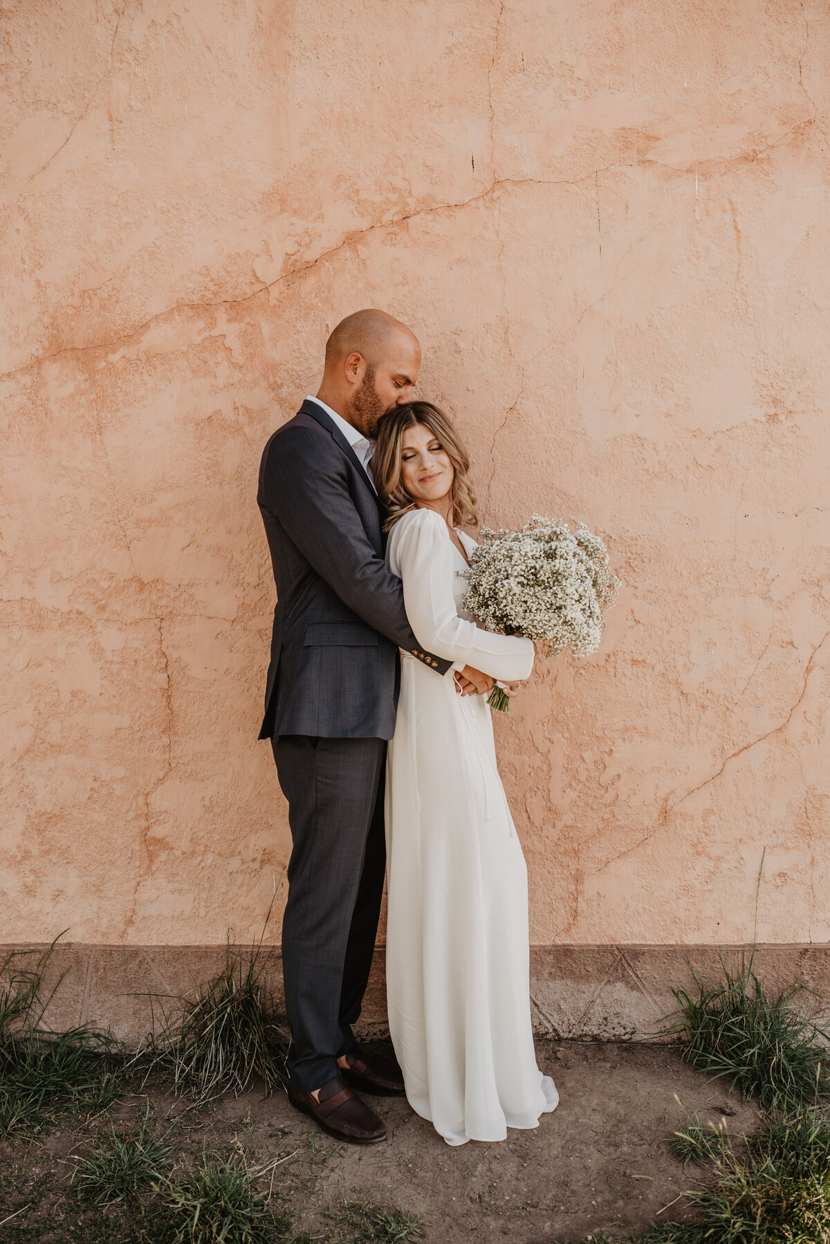 jackson wyoming photographer photographs wyoming elopement bride and groom standing by a terracotta wall with the groom holding the bride while standing behind her as she holds her babies breathe wedding bouquet and the groom kisses her head