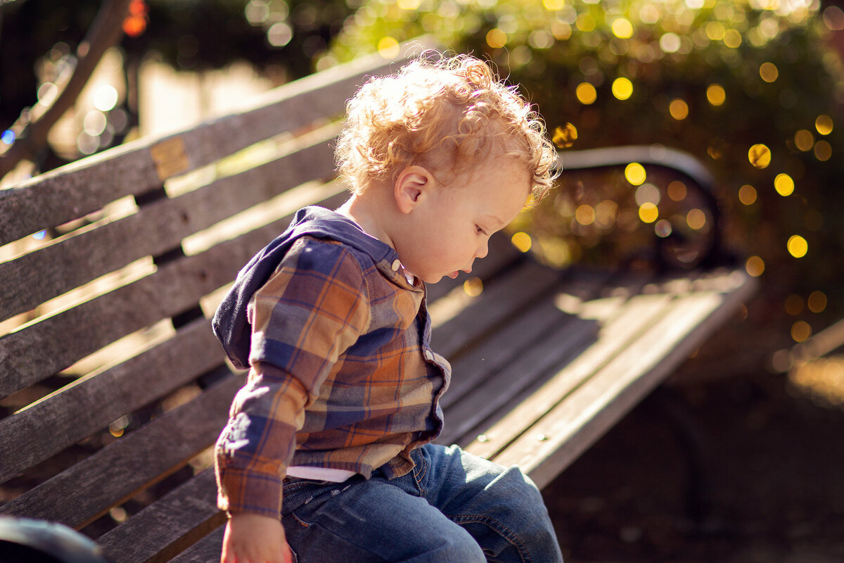 Curly haired toddler is sitting on a bench in Carmel, Indiana.