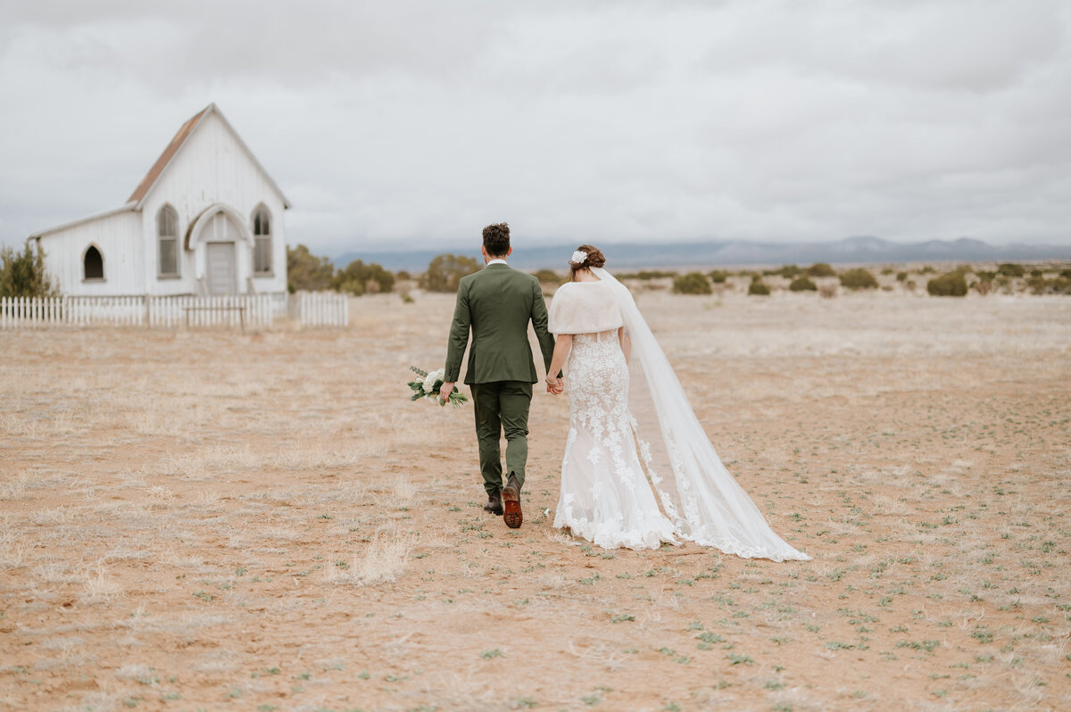 bride and groom holding hands walking in a field with a white chapel