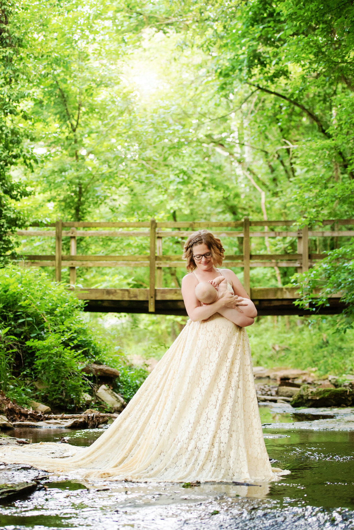 st-louis-motherhood-photographer-mother-in-lace-gown-cradling-baby-standing-in-creek-with-greenery-and-bridge-in-background