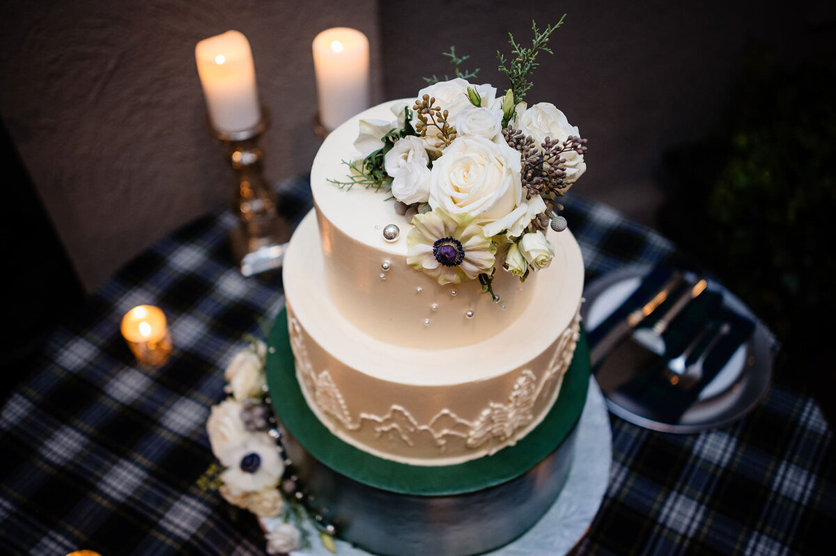 An elegant two-tiered wedding cake with a gold base layer and a white top layer, adorned with white flowers and greenery, on a plaid tablecloth next to lit candles.