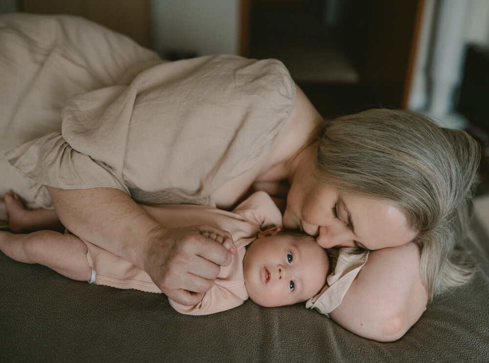 Laid-back newborn portrait of mother cuddling and kissing baby while lying on bed, capturing real-life love and comfort in an organic lifestyle session.