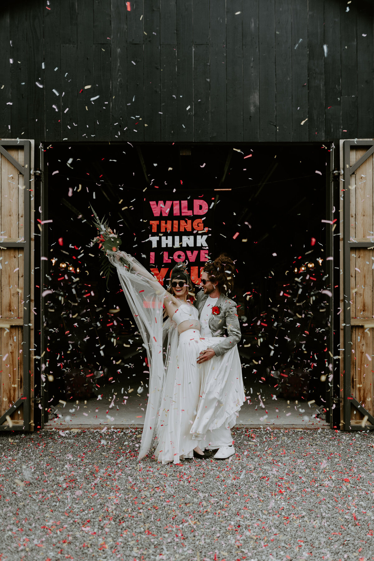 A cool couple stand outside The Giraffe Shed, an alternative barn wedding venue in mid-Wales. They are being showered in confetti in front of a custom wedding banner that says Wild Thing, I Think I Love You.