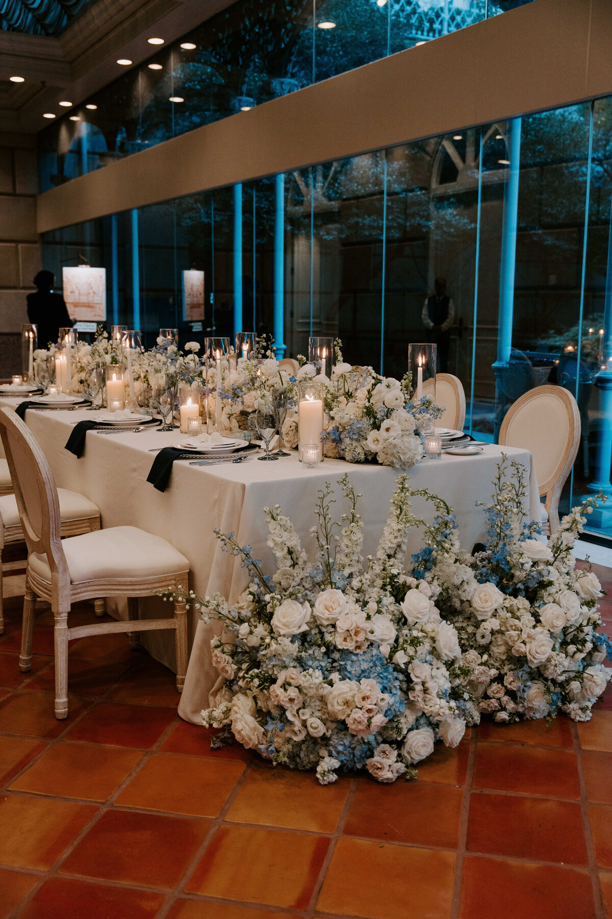 Elegant wedding table setup with white linens, surrounded by cream chairs and adorned with lush white and blue floral arrangements and glowing candles.