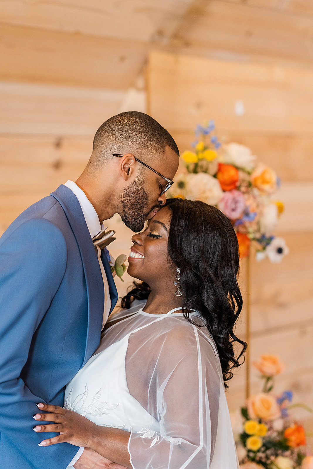 Black couple embracing , groom kissing forehead, colorful flowers in the backround