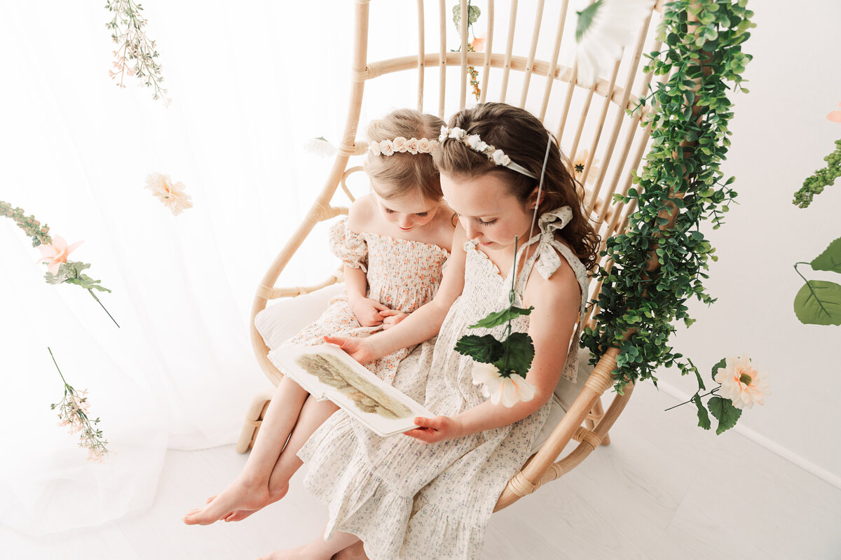 Two young girls in floral dresses sit together in a hanging rattan chair, immersed in a storybook, surrounded by gentle touches of greenery and flowers, creating a serene and whimsical atmosphere.