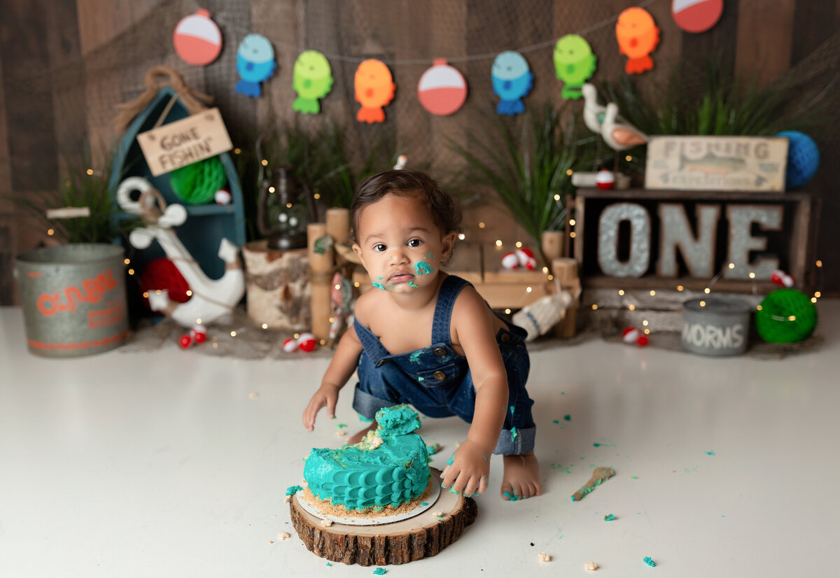 Fishing themed cake smash at West Palm Beach photography studio. Baby boy has smashed teal-colored cake wearing overalls and looking at the camera. In the background, there is a fishing pennant, anchors, boats, buoys, and buckets.