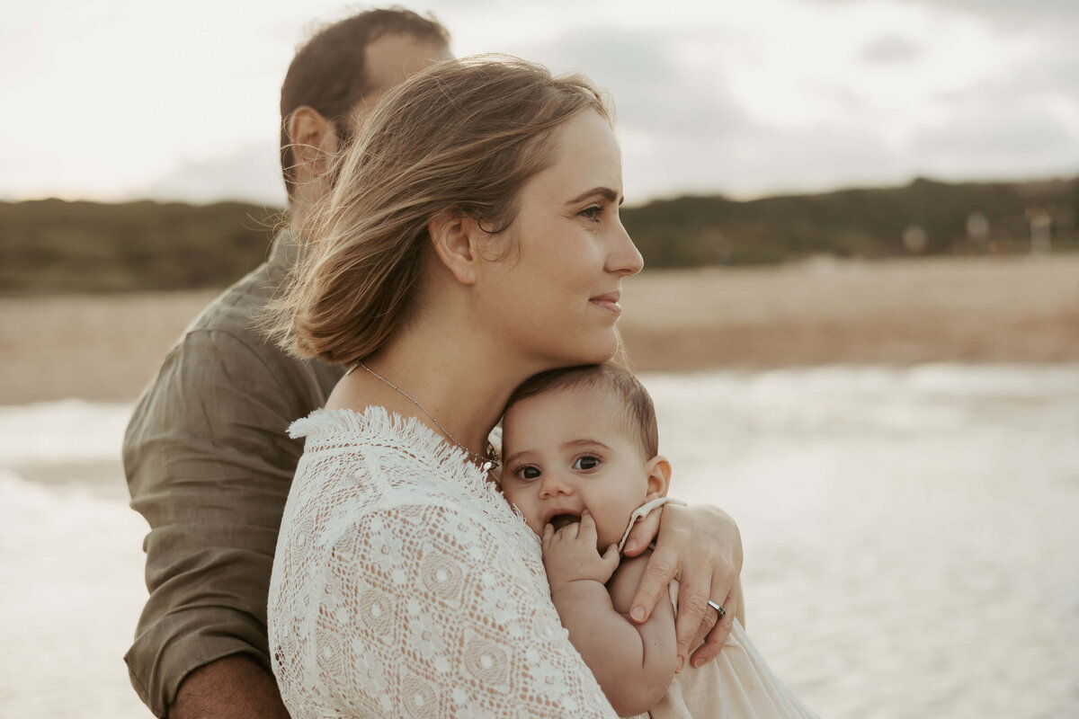 Mum and dad staring out to sea while baby has his  fingers in his mouth during their photoshoot at a Sydney beach.