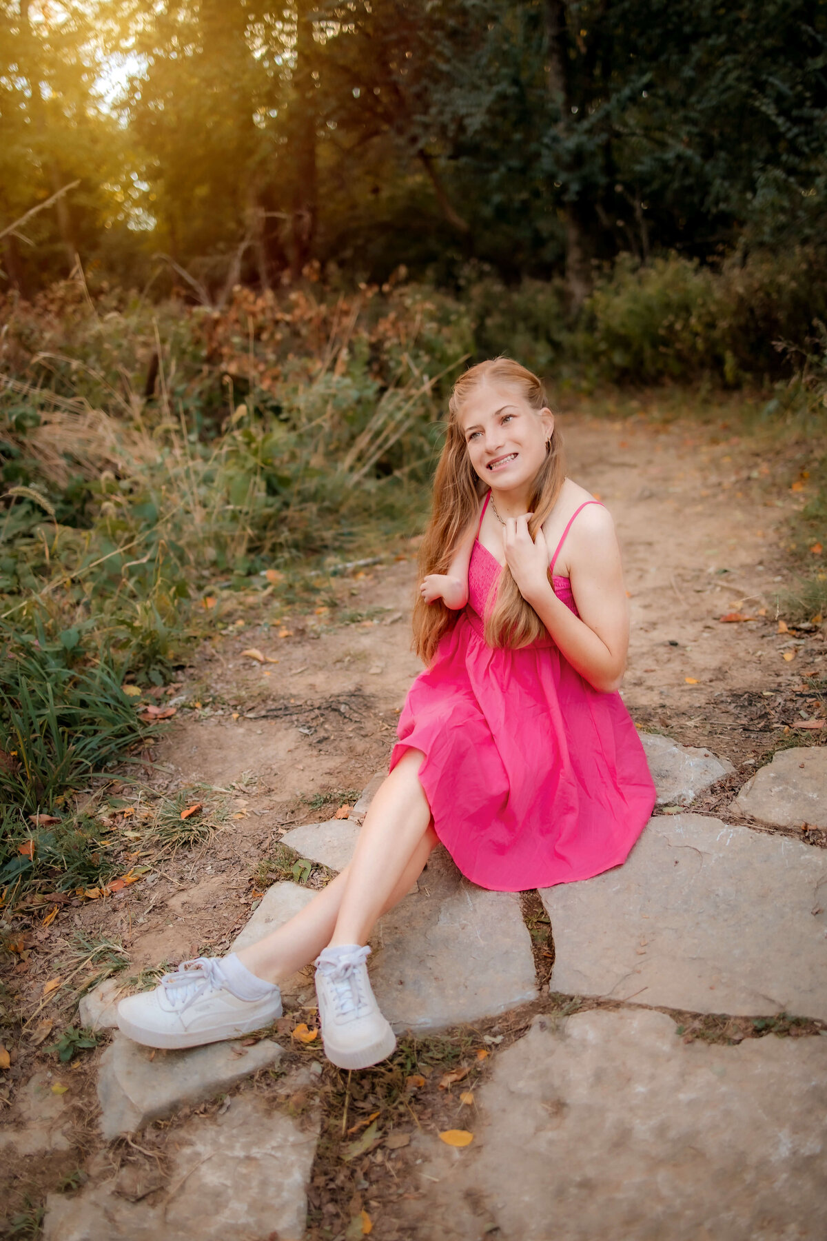 A beautiful long blonde haired girl is sitting on a natural stone trail in a pink summer dress.