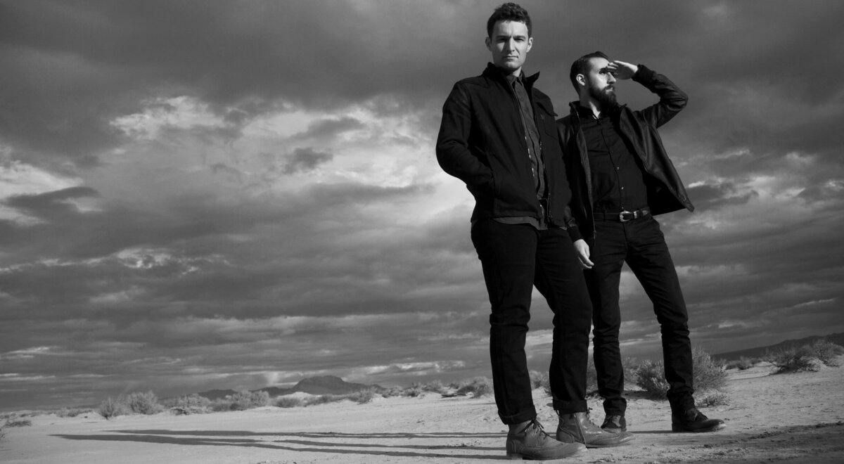 Black and white band portrait Polarcode duo standing in desert at sunrise one member shielding eyes with hand El Mirage