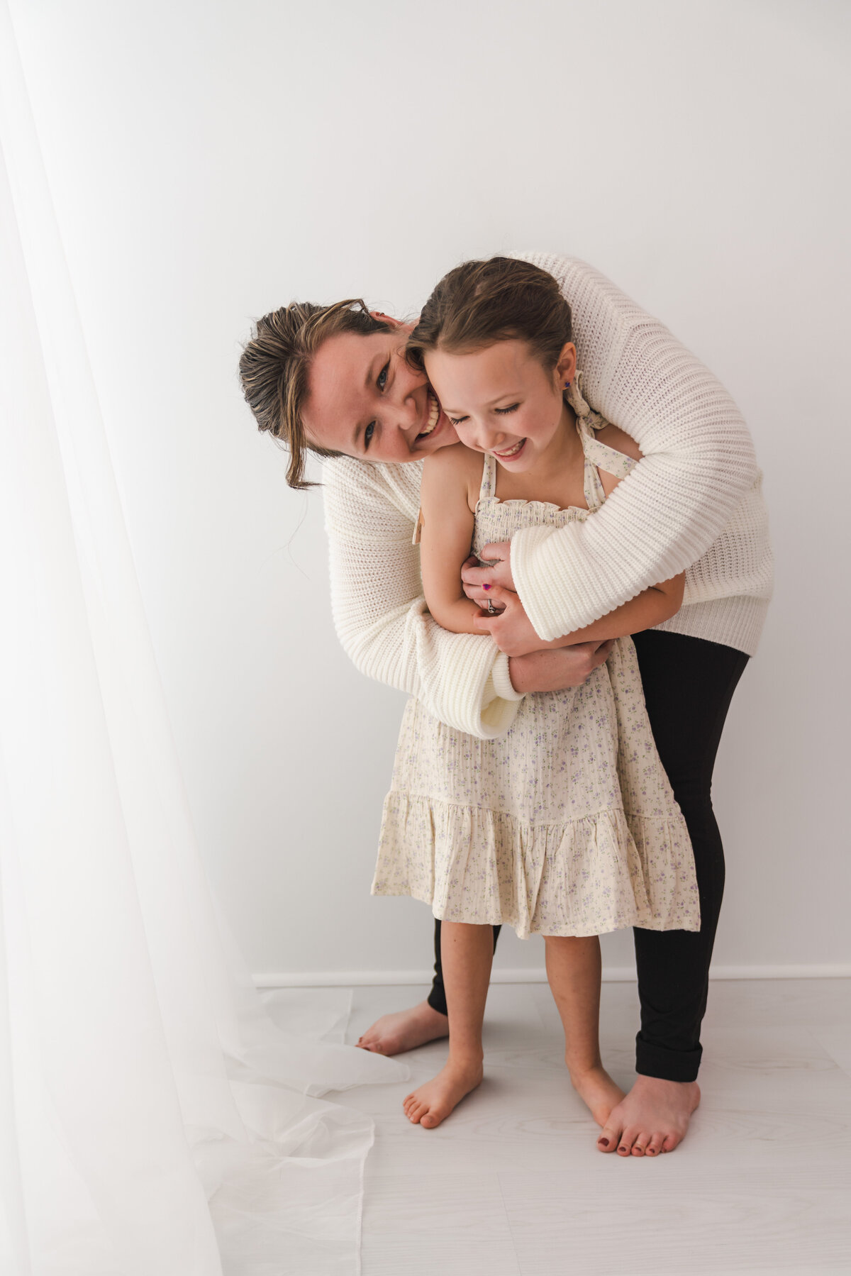 A heartwarming moment between a smiling woman and a giggling young girl as they share a close embrace in a bright, minimalist room.
