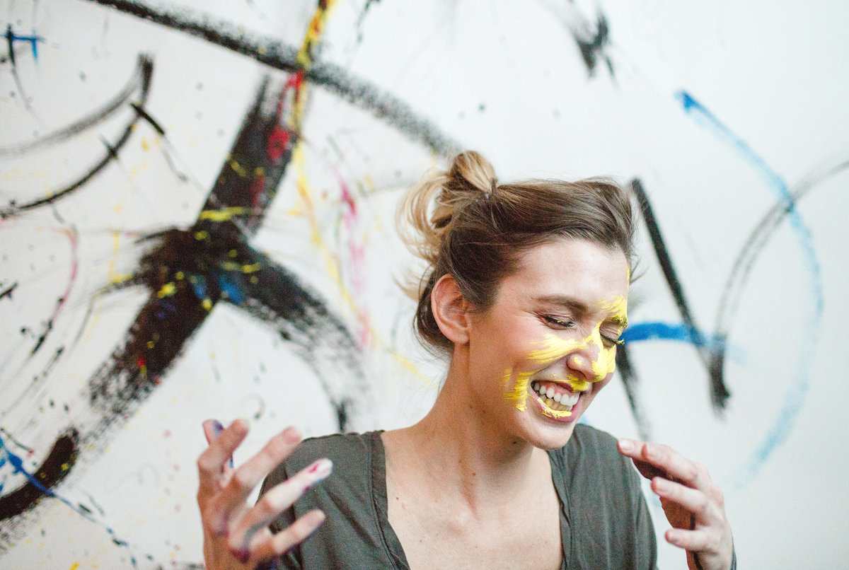 anna canfield laughs after she paints her face with a paintbrush of eyllow paint against her artwork background
