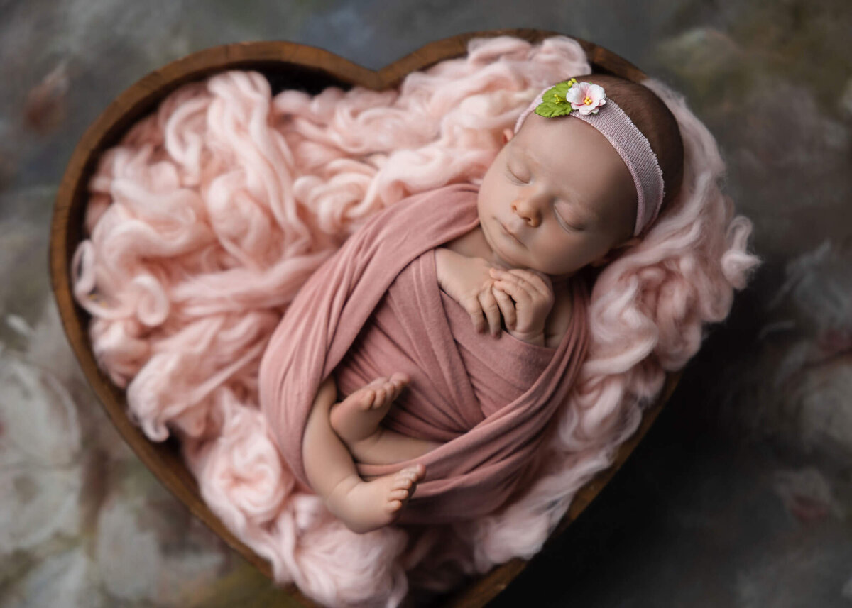 newborn baby wrapped in pink fabric with a pink headband asleep in a pink heart shaped bowl