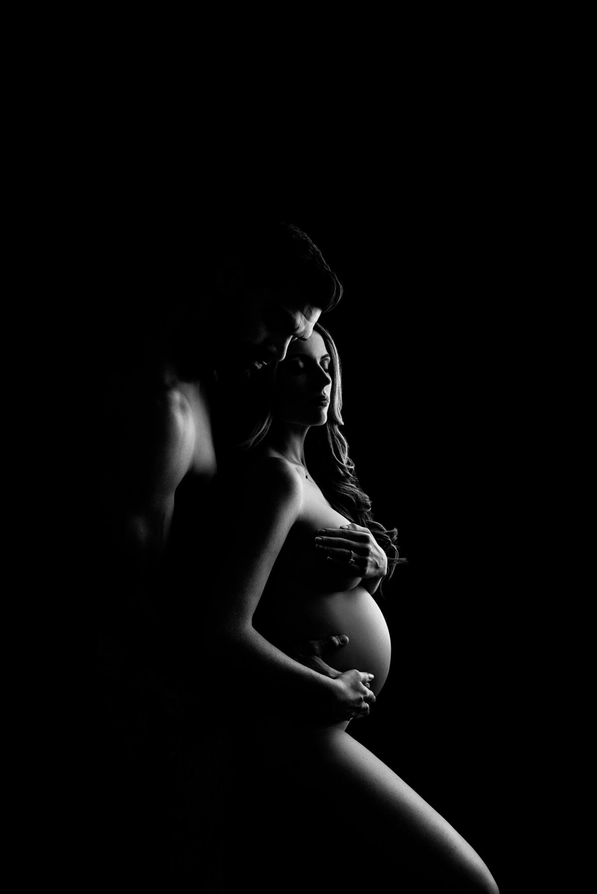 An intimate fine art maternity photoshoot captured by Katie Marshall, best maternity photographer in New Jersey. A black and white images features an expectant mom standing side bare and side profile to the camera with her husband standing behind her. His cheek is resting against her temple and both have their eyes closed. The woman's forearm is covering her breasts and her husband's hand is resting underneath her bump. There are alot of highlights and shadows to add great dimension to the photo.