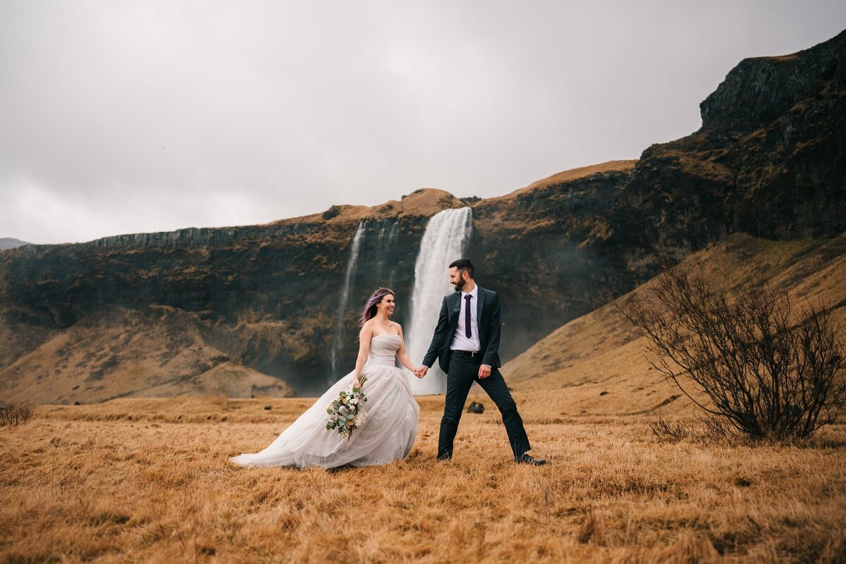 Hand in hand, smiles exchanged, this couple walks with joy, framed by the majestic backdrop of a waterfall in Iceland. A moment of pure happiness on their special day.