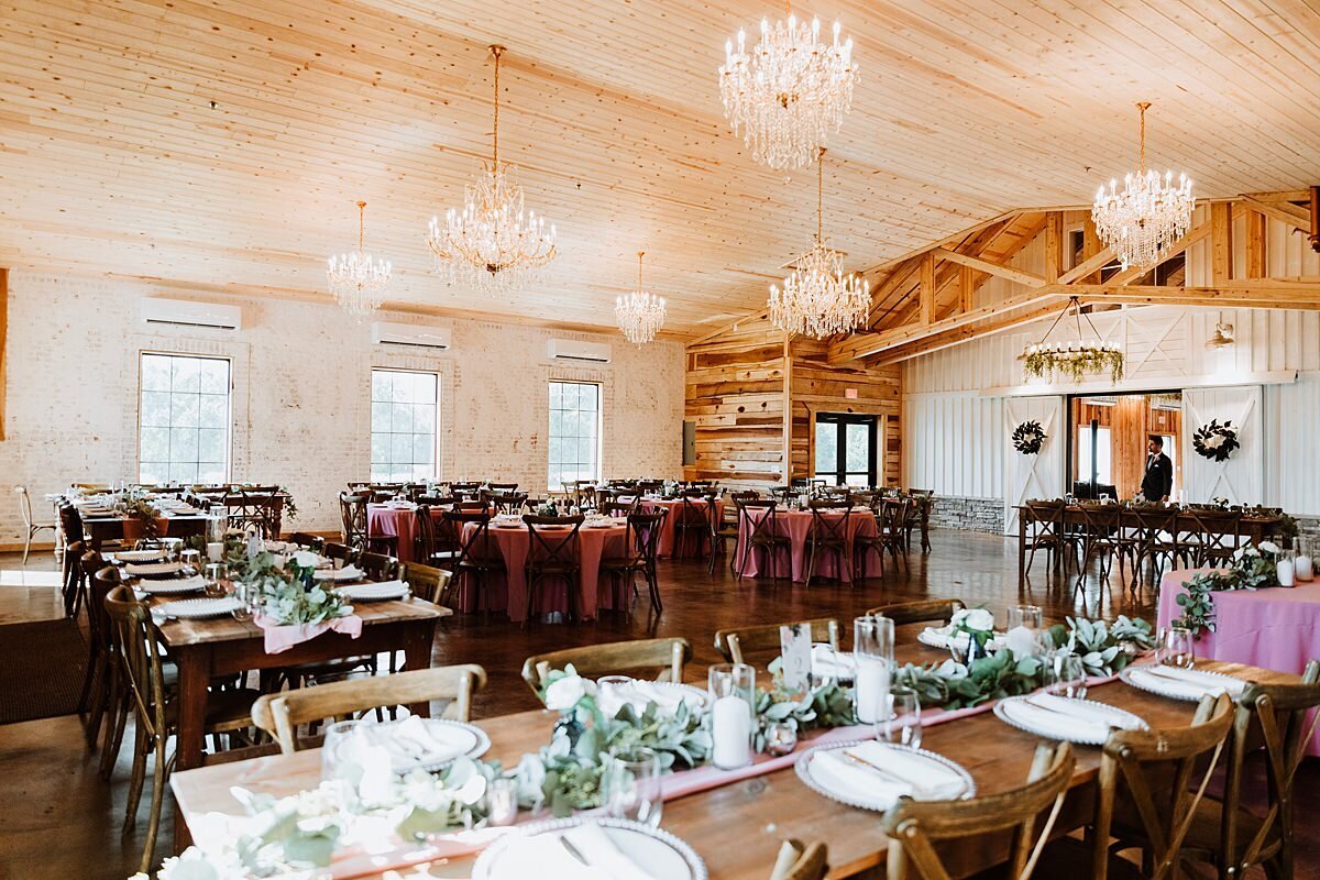 Brown farm tables set with blush table runners and long garlands of greenery and blush roses. Glass cylinders with white pillar candles, silver chargers and white plates are at each place setting. Crystal chandeliers hang from the wood plank ceiling at Steel Magnolia Barn.