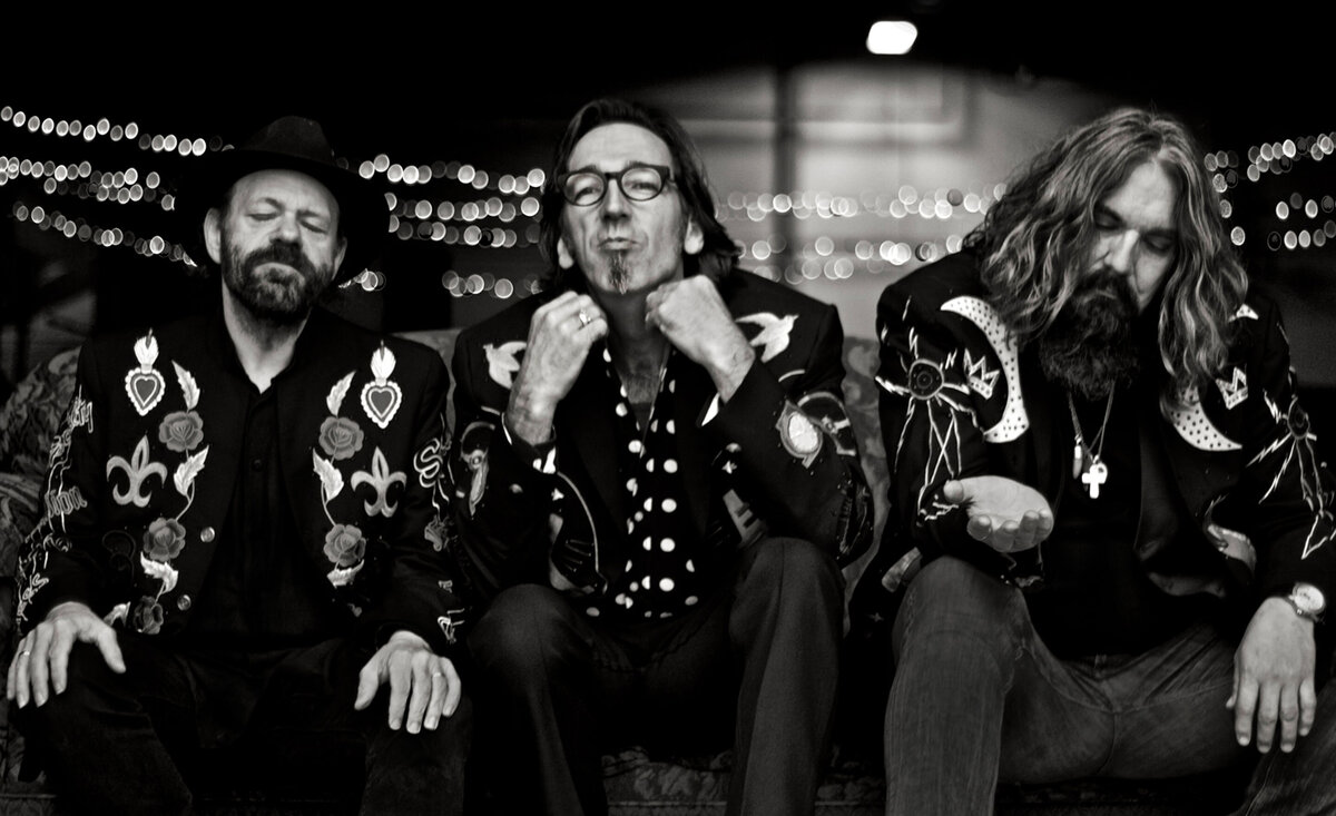 Musical trio portrait Blackie And The Rodeo Kings black and white sitting close together wearing patterned suit jackets middle member adjusting collar lights hung behind