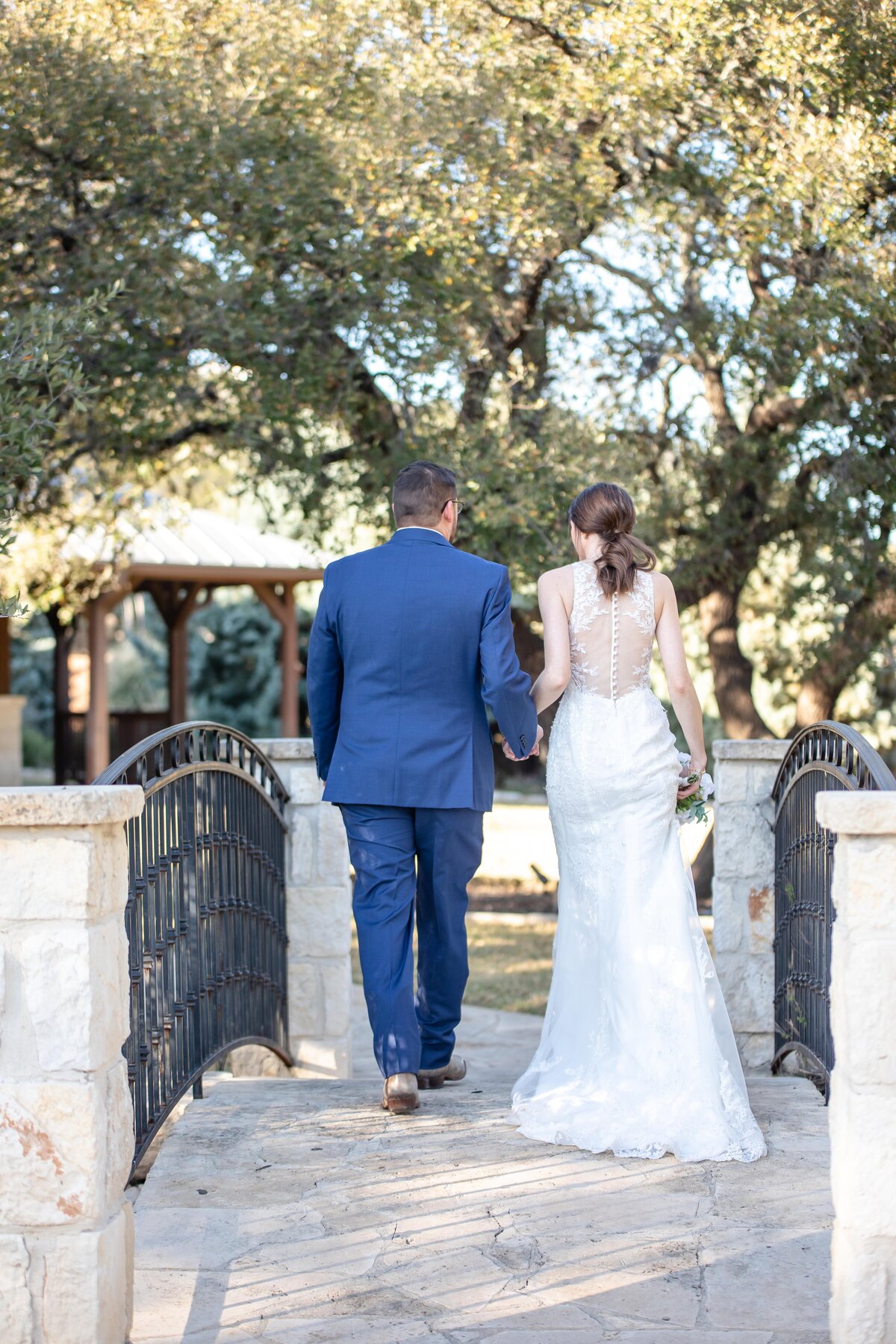 bride and groom in navy suit walk away from photographer on bridge leaving Boerne Texas ceremony
