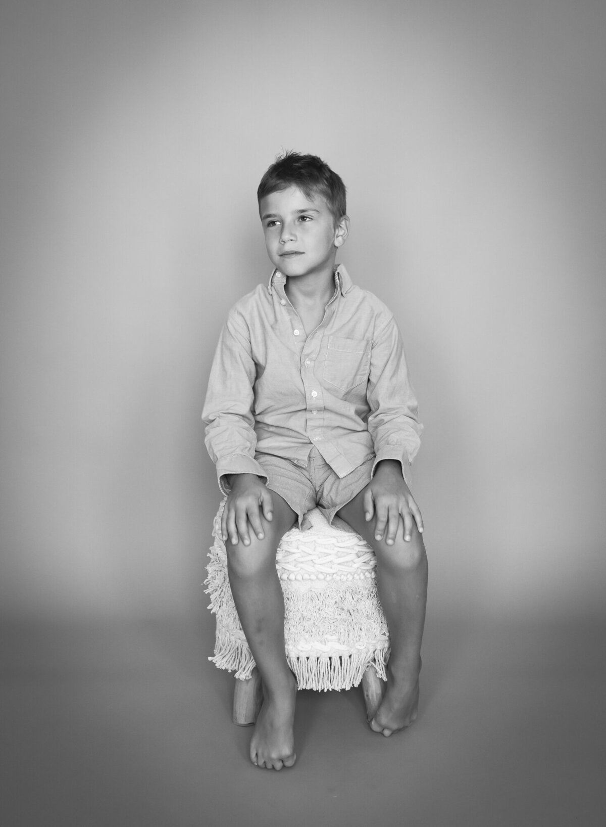 beautiful black and white  soft edit portrait of barefoot boy in dress shirt  sitting on a boho stoll, looking away
