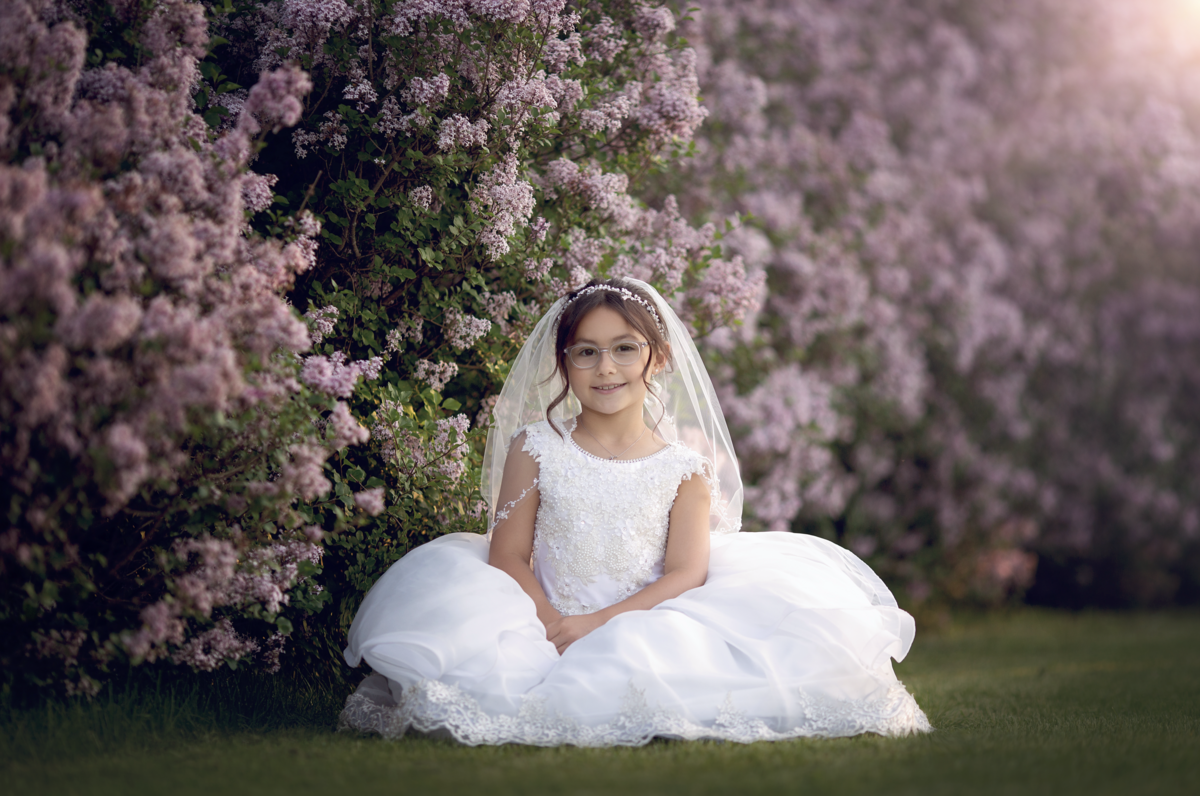 A girl in a large while dress and veil sits in a park with pink flowering bushes at sunset taken by a New Jersey Communion Portrait Photographer
