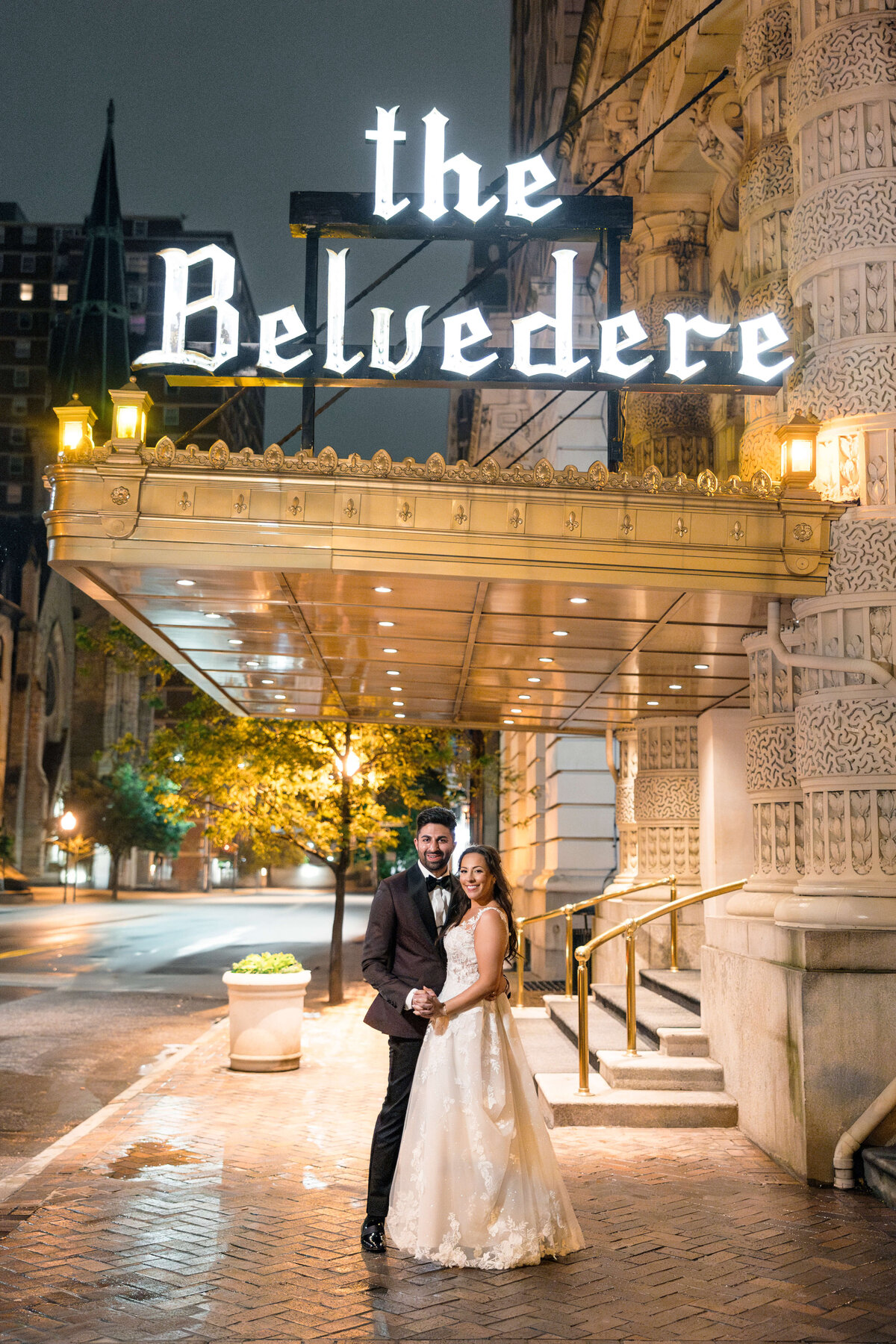 A couple in wedding attire poses under the illuminated sign of The Belvedere on a quiet city street at night. The bride wears a white gown, and the groom is in a dark suit. The entrance is elegant with ornate details and warm lighting.