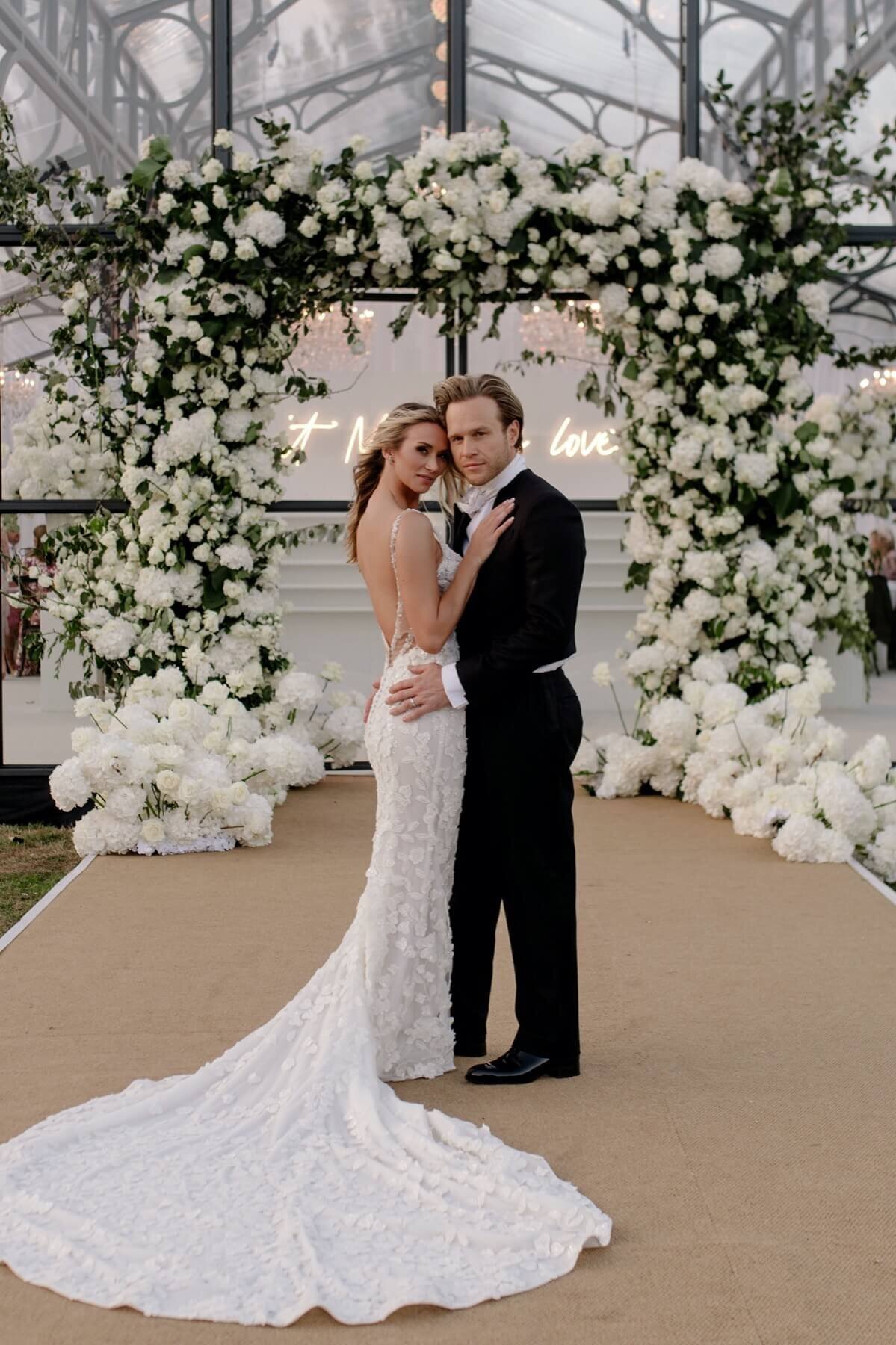 Amelia and Olly Murs in front of their glass wedding marquee with a stunning floral arch entrance.