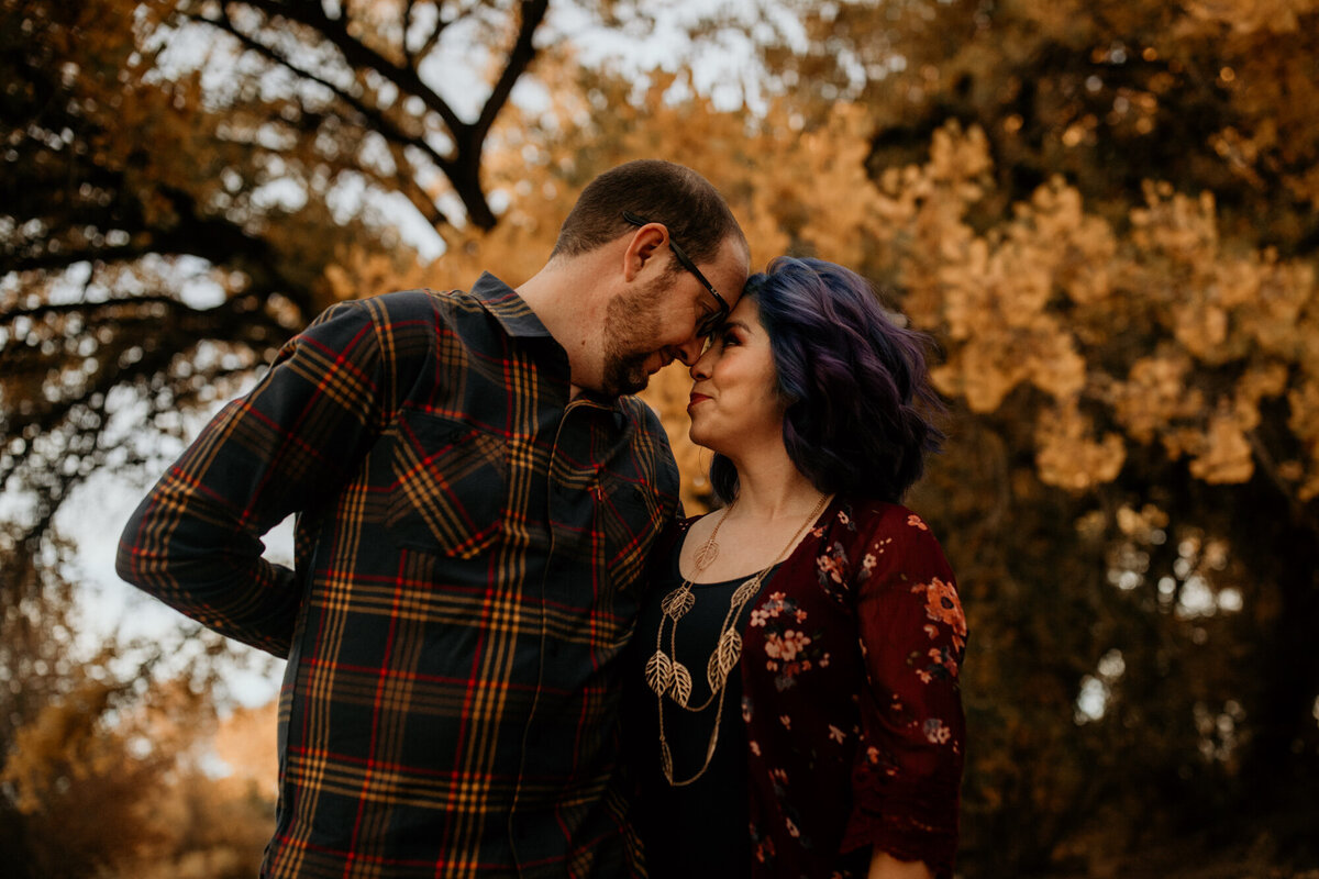 husband and wife with their foreheads together and fall trees behind them