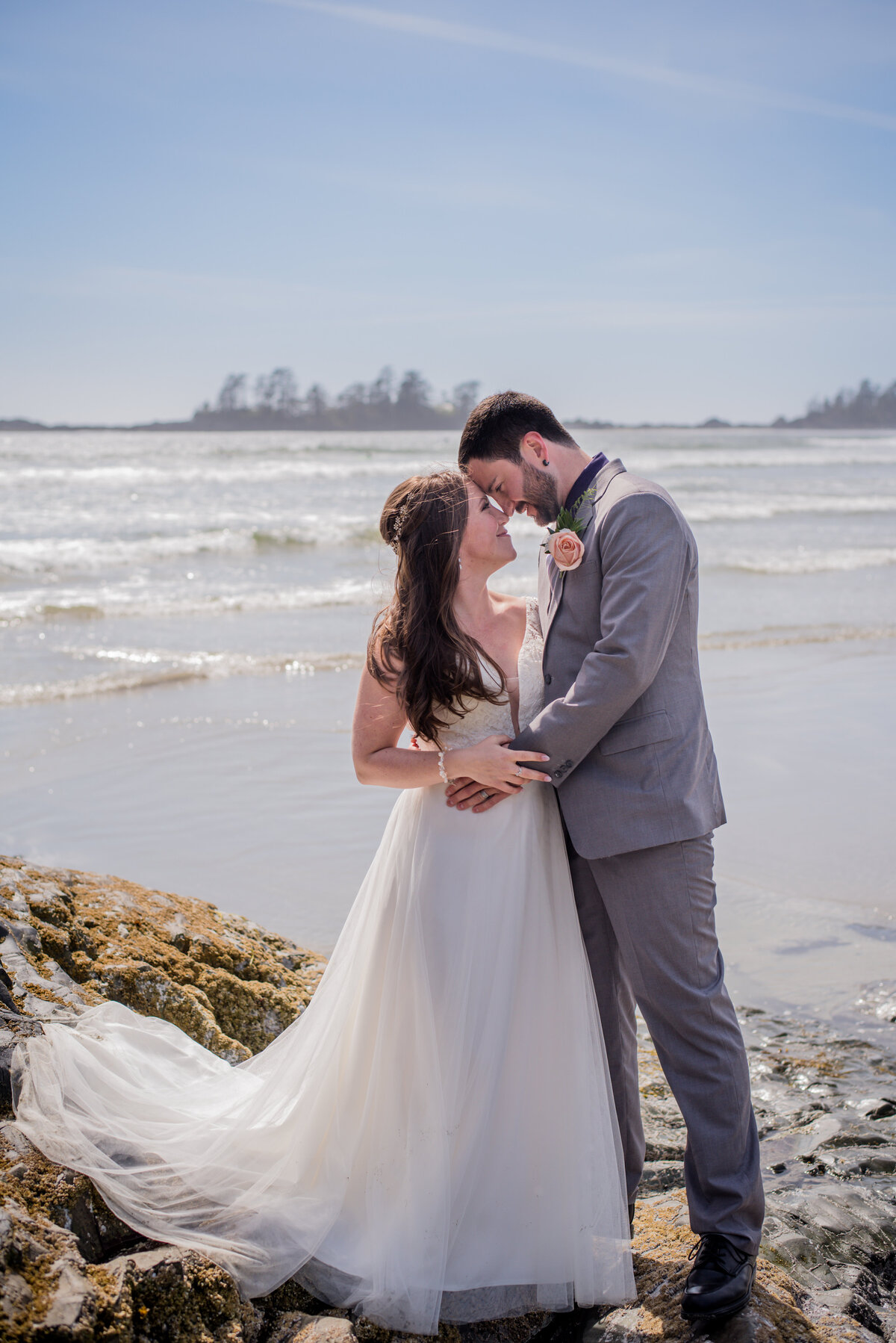 Bride and groom stand on a big rock foreheads touching closely, behind them is the ocean in Tofino BC.