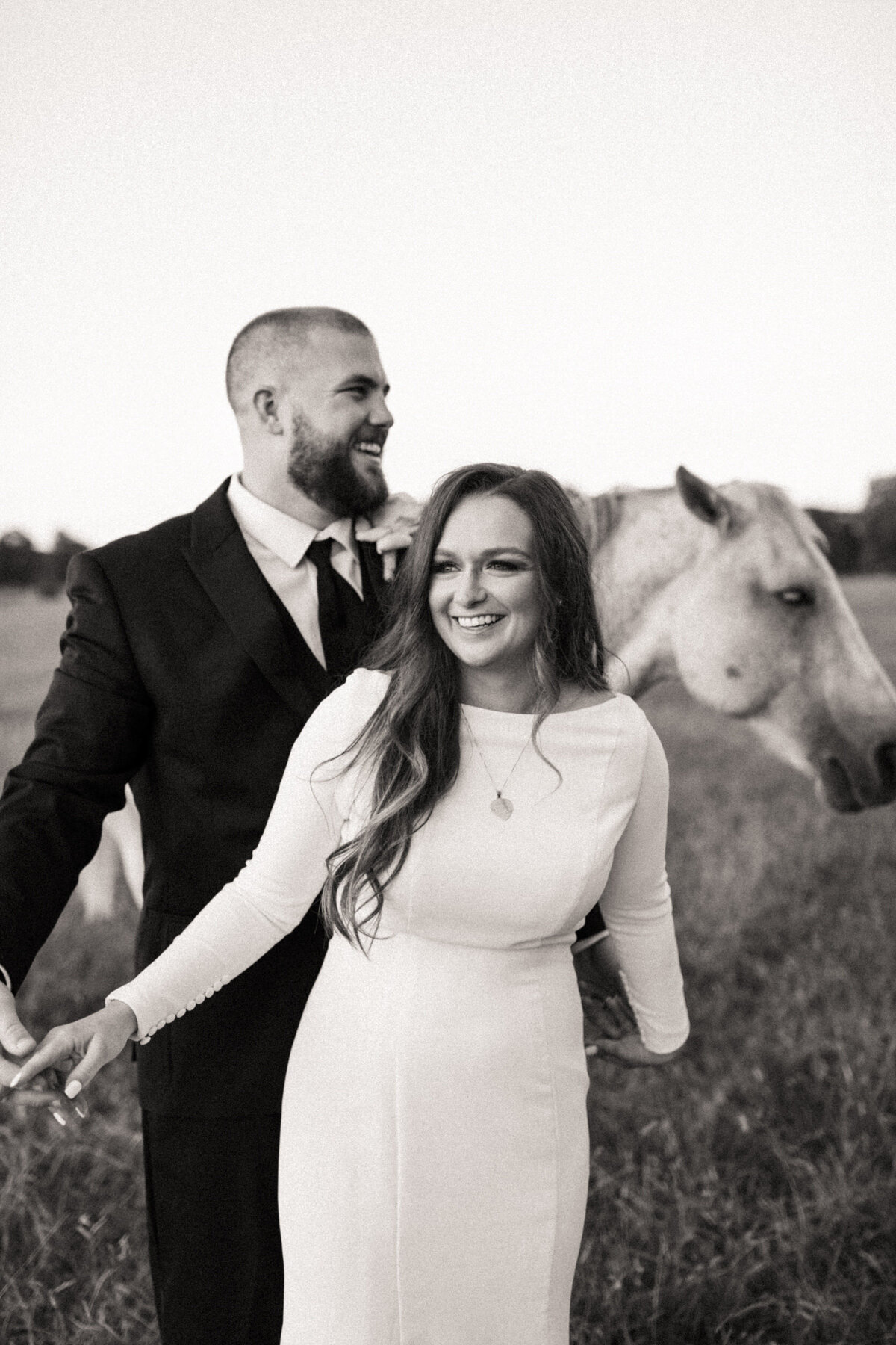 Bride and groom laughing in a field with a horse behind them