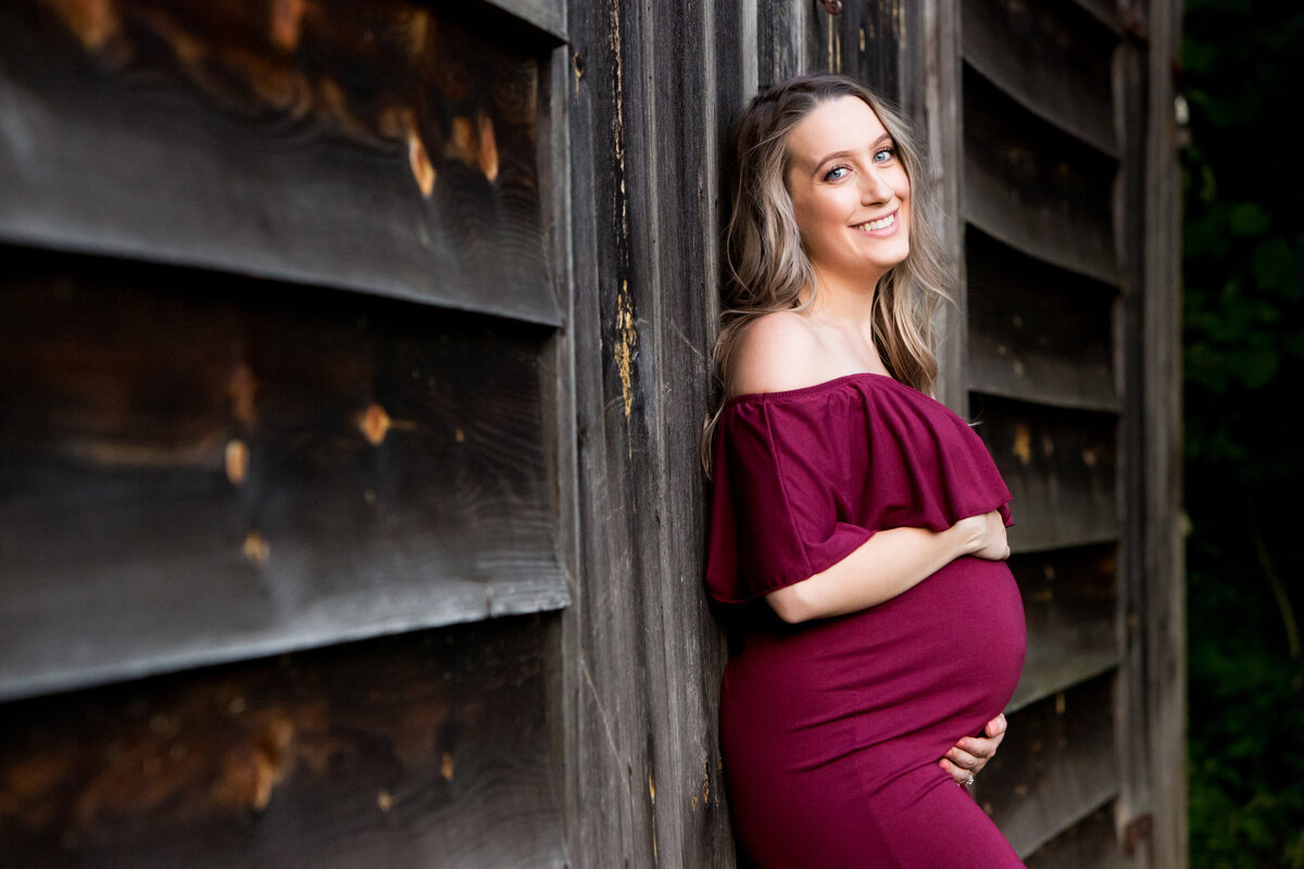A pregnant white woman in a maroon maternity dress leans against dark wood New City maternity photographer Milestone Images