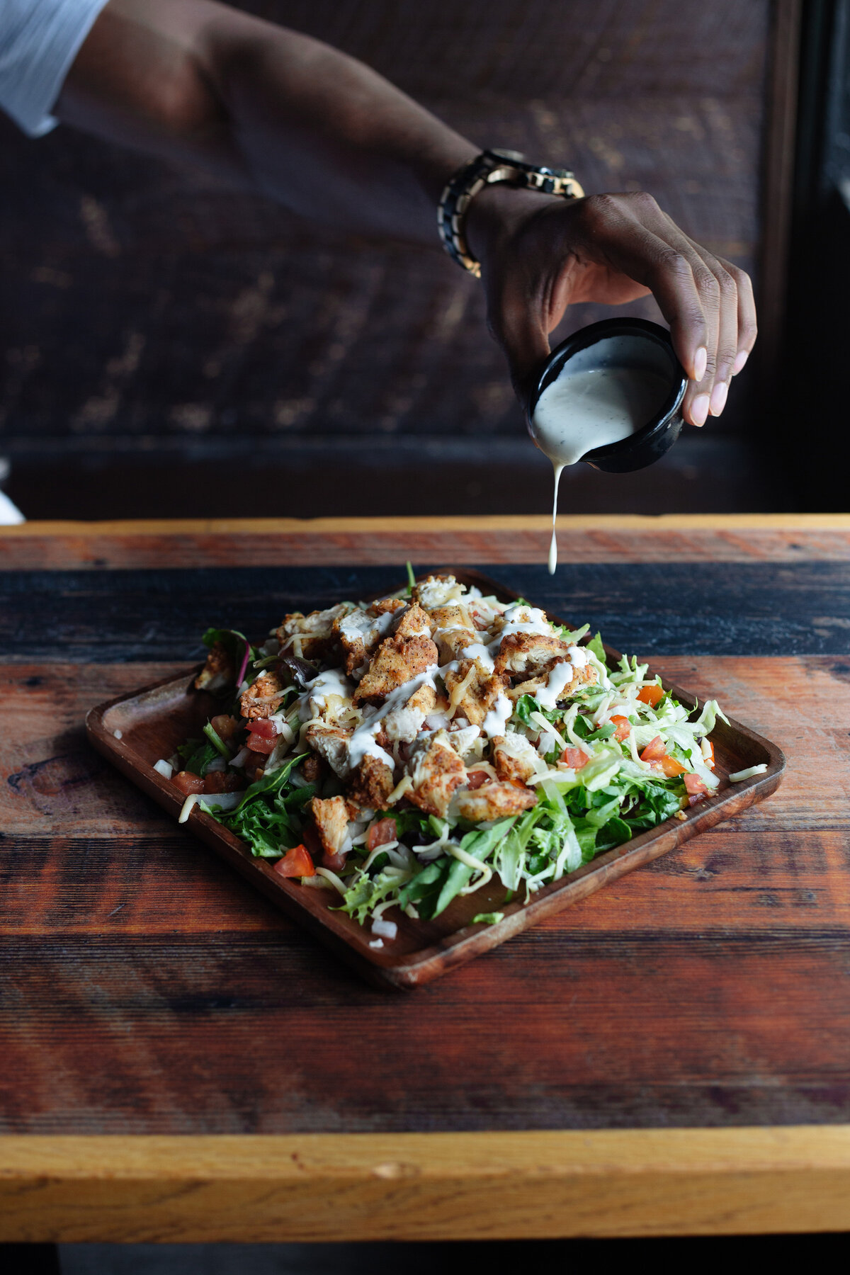 Ranch dressing being poured over a fried chicken salad on a wooden plate set on a wooden table. Natural light food photography.