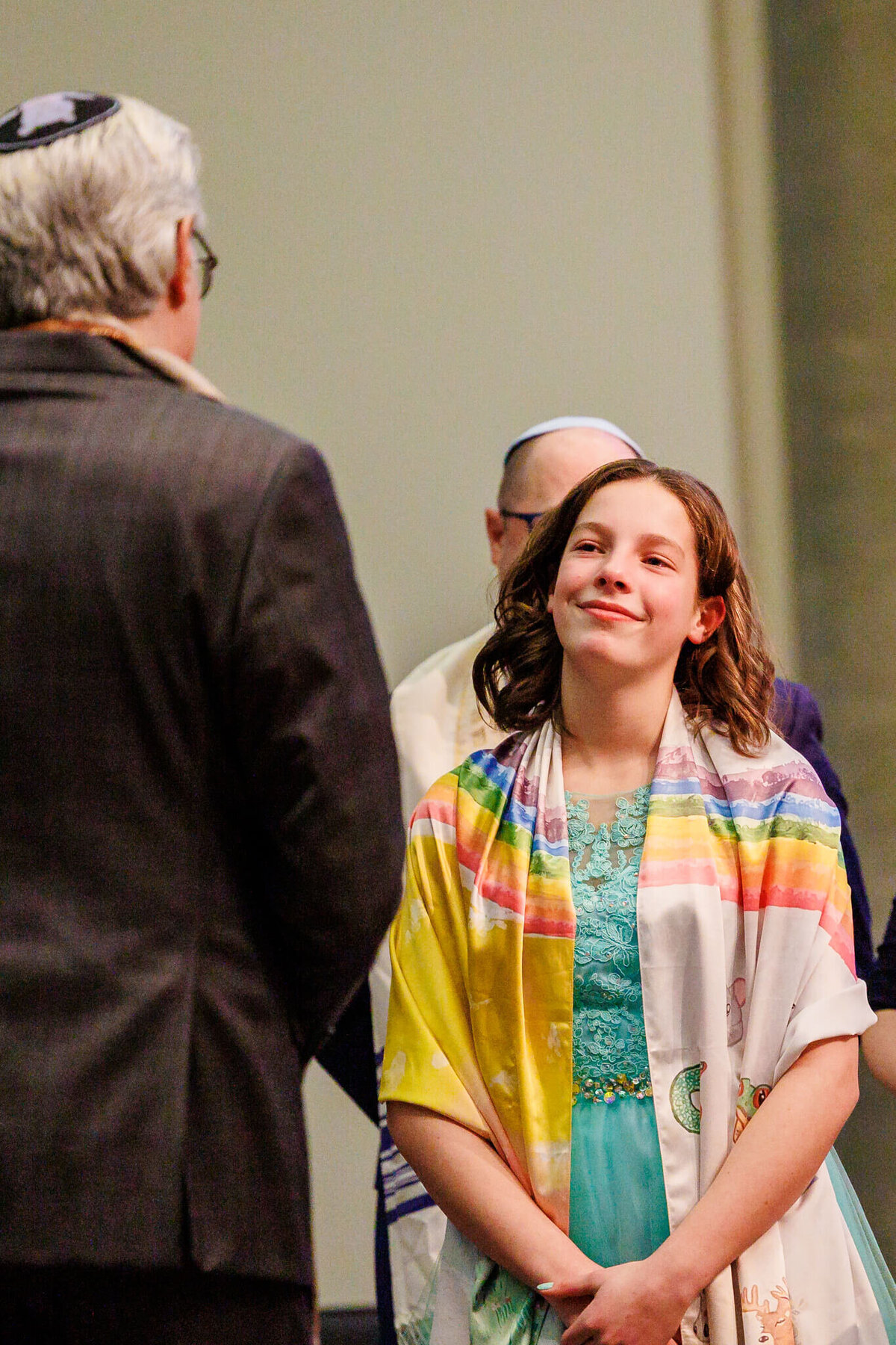 A teenage girl listens with a smile to her rabbi while wearing a blue dress under a rainbow tallit