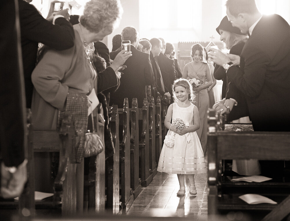 flower girl wearing short dress walking up the aisle of the church while wedding guests look on