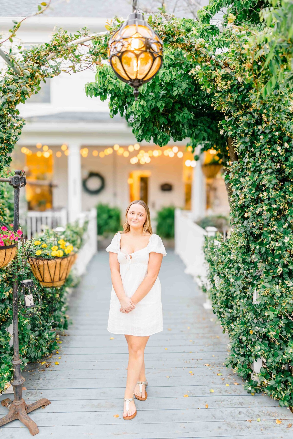A woman in a white dress stands gracefully on a wooden path adorned with greenery and hanging flower baskets. She's captured in front of a charming white house with porch string lights by a talented senior portrait photographer from Fayetteville, NC. A decorative light fixture completes the scene.