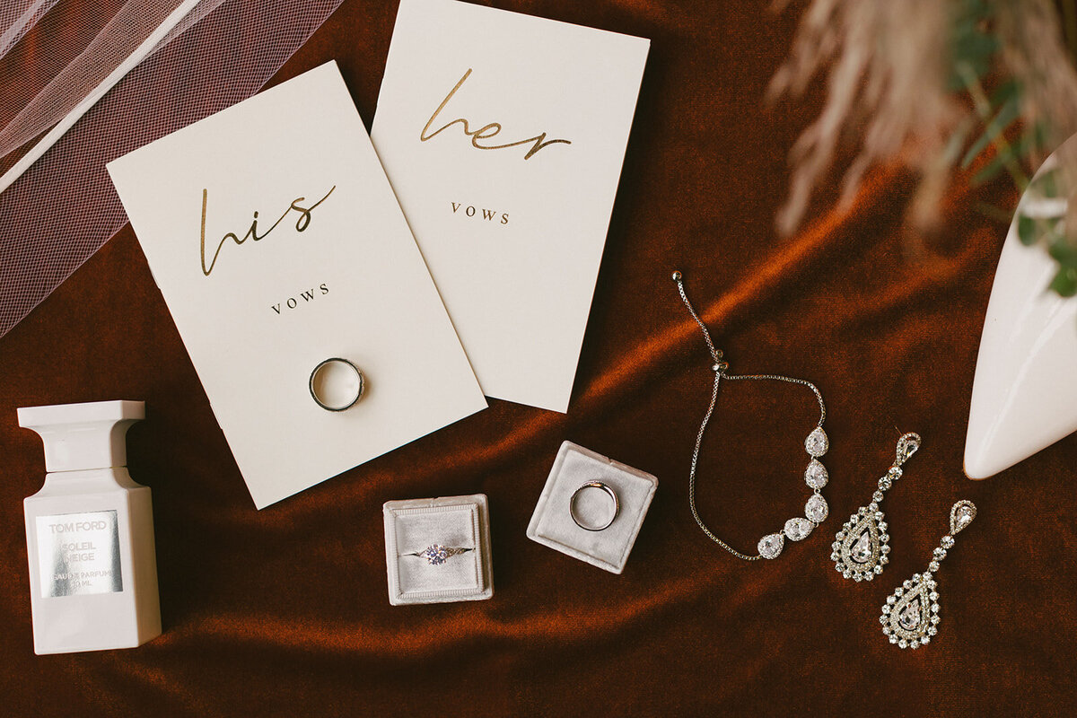 Elegant jewelry, wedding bands and vows lay on lush red velvet for a flat lay photo displaying timeless wedding details at Loft on Lake.