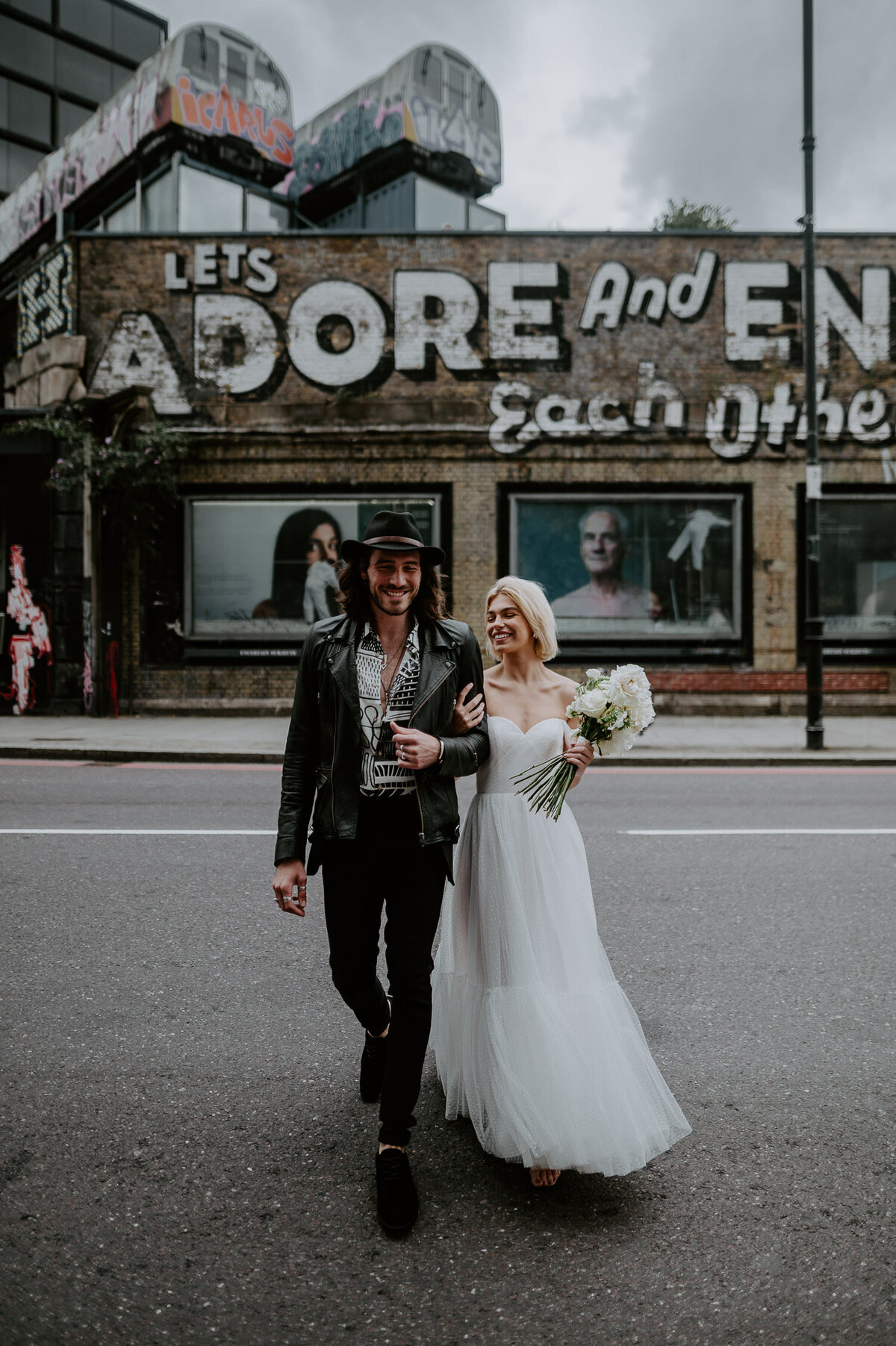 An alternative couple cross the road in Shoreditch, London. The bride is wearing a shoulder cut Justin Alexander wedding dress whilst the groom is dressed casually with a black hat and boots.