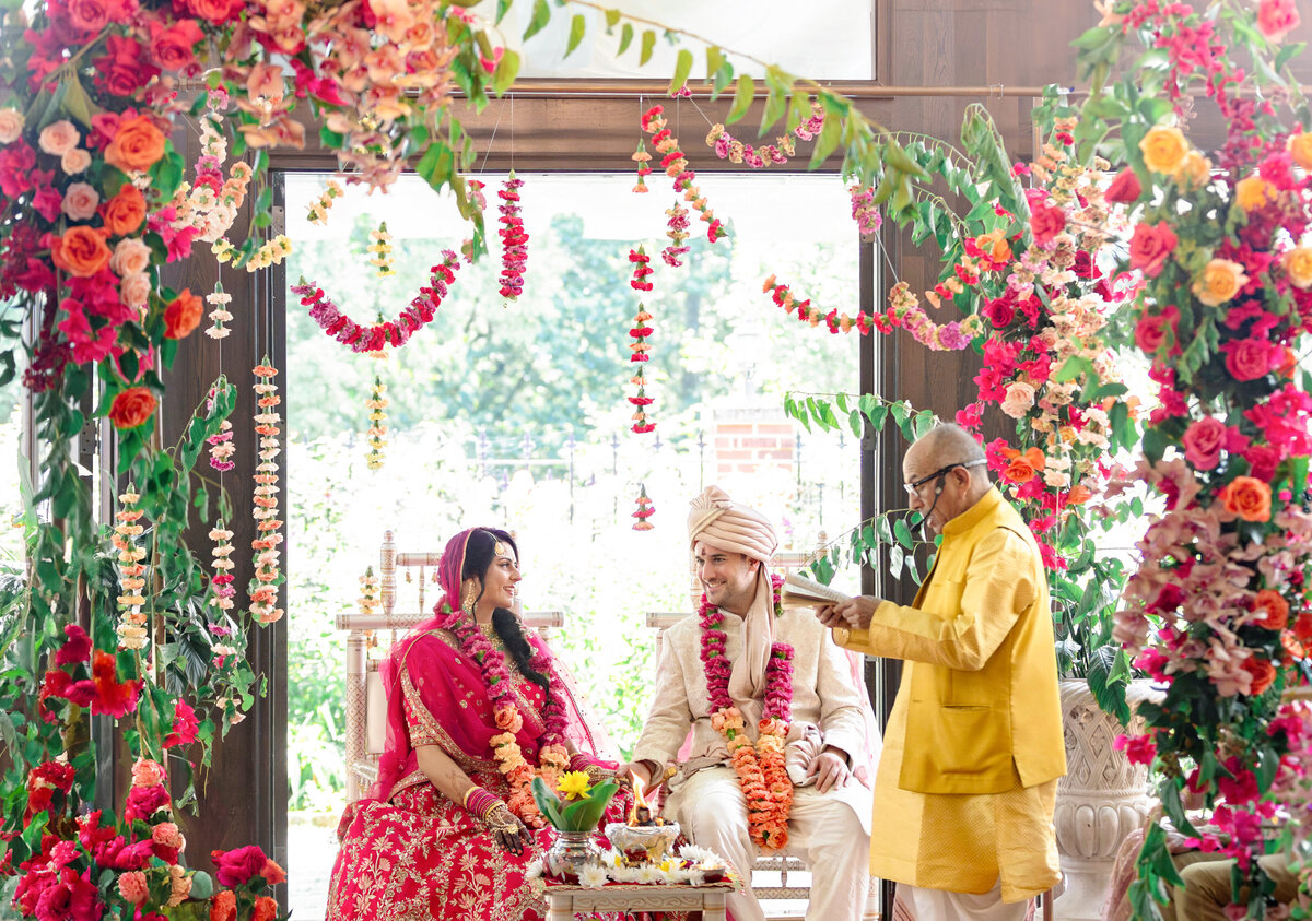 A couple in traditional attire sits under a vibrant floral canopy during a wedding ceremony. The man wears a white sherwani, and the woman wears a red saree. A priest in yellow officiates, surrounded by lush flowers and garlands.