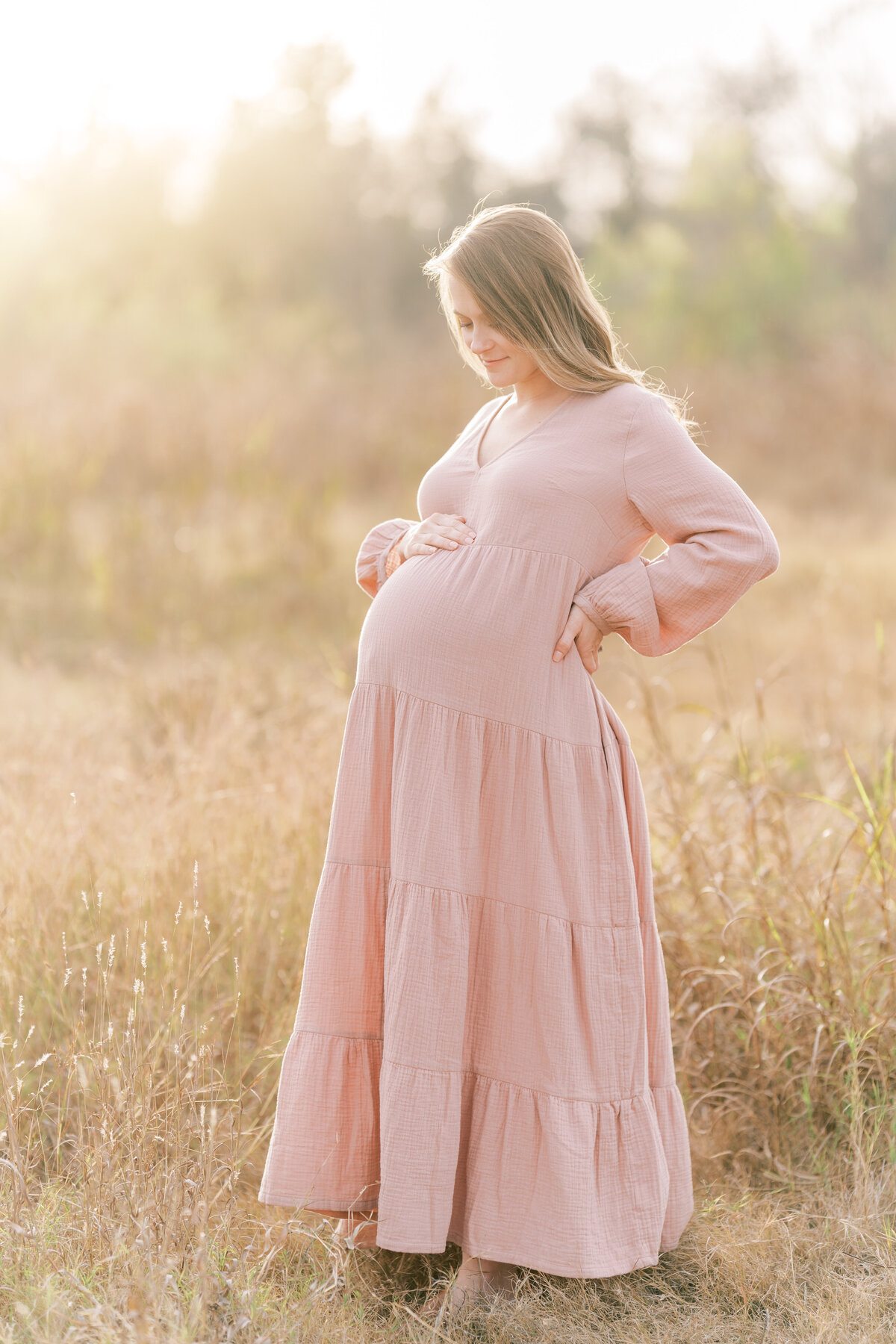 Portrait of a pregnant woman in a country field wearing a pink sundress, smiling while looking down at her belly.