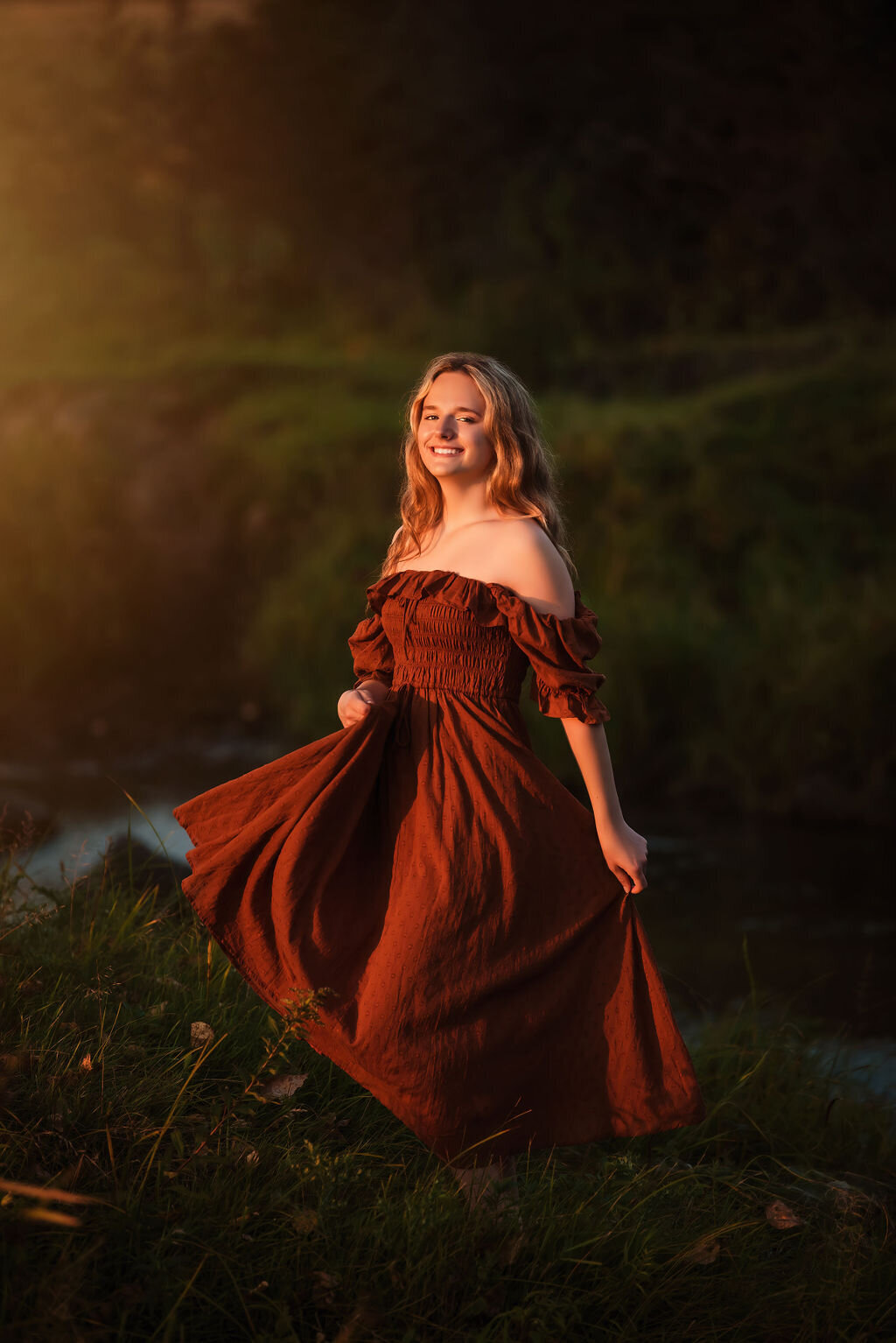 A young woman in an orange flowing dress dancing in a field in front of a river at sunset, with warm light illuminating her joyful expression and flowing hair, captured by a Bemidiji, Minnesota fine art senior photographer