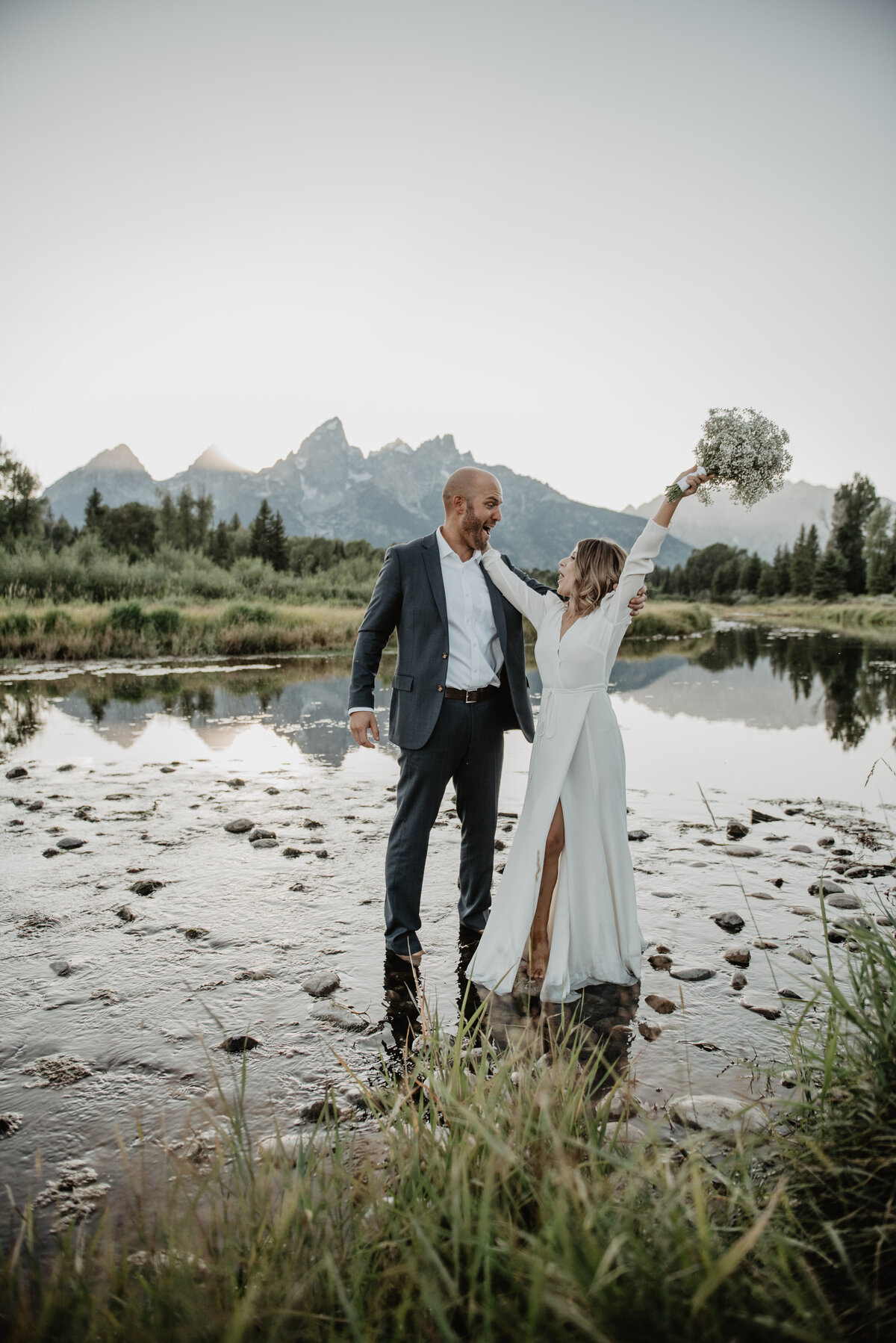 jackson wyoming photographer captures Bride and groom in a creek in the Tetons celebrating their elopement, man smiling at his wife and woman with her arms in the air with her white bouquet