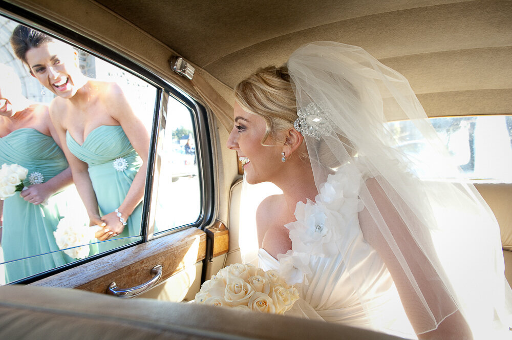 bride with blonde hair wearing a one strap, mermaid style wedding dress looking out the window of a vintage car at her bridesmaids wearing sage green, column style dresses
