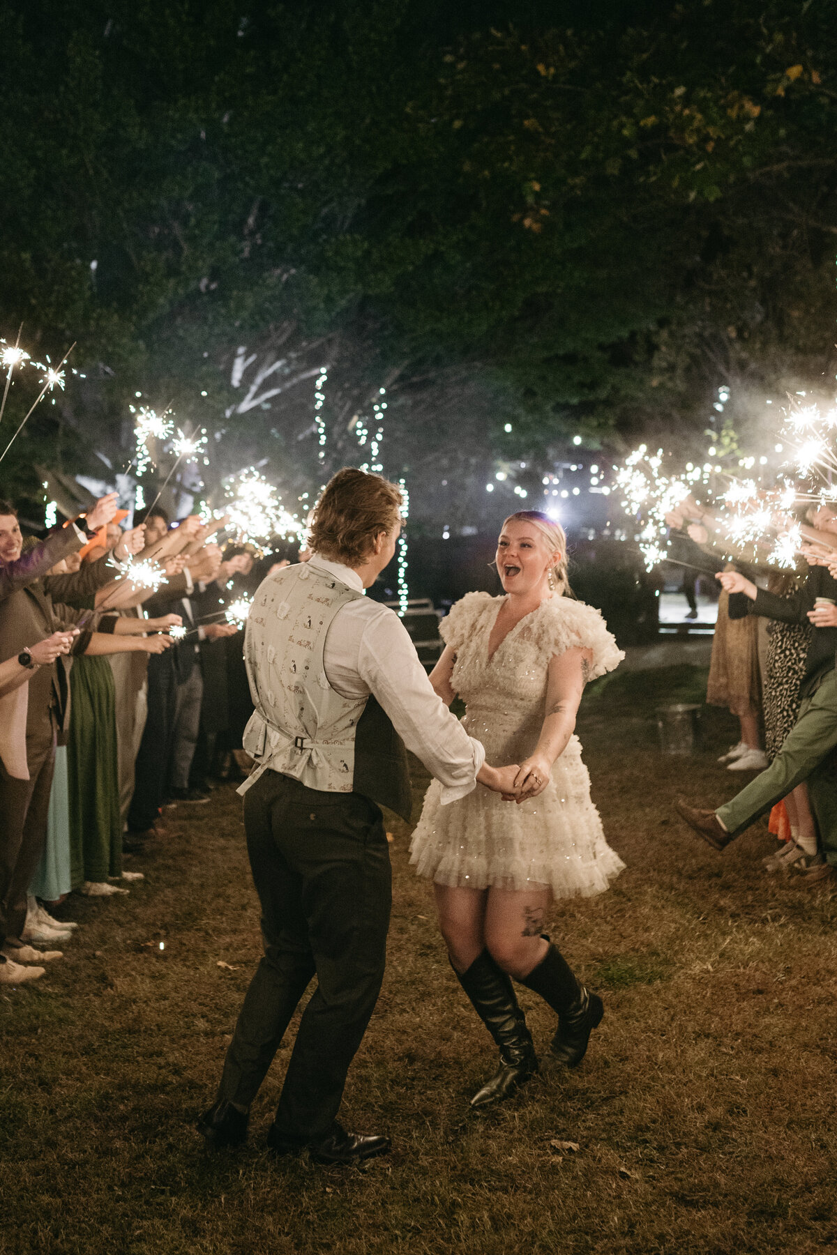 Bride and groom dancing joyfully during nighttime sparkler exit surrounded by cheering guests at The Nest in Northwest Arkansas. Bride wearing a short ruffled reception dress and boots, celebrating with a whimsical, storybook-style wedding atmosphere enhanced by garden-inspired florals.