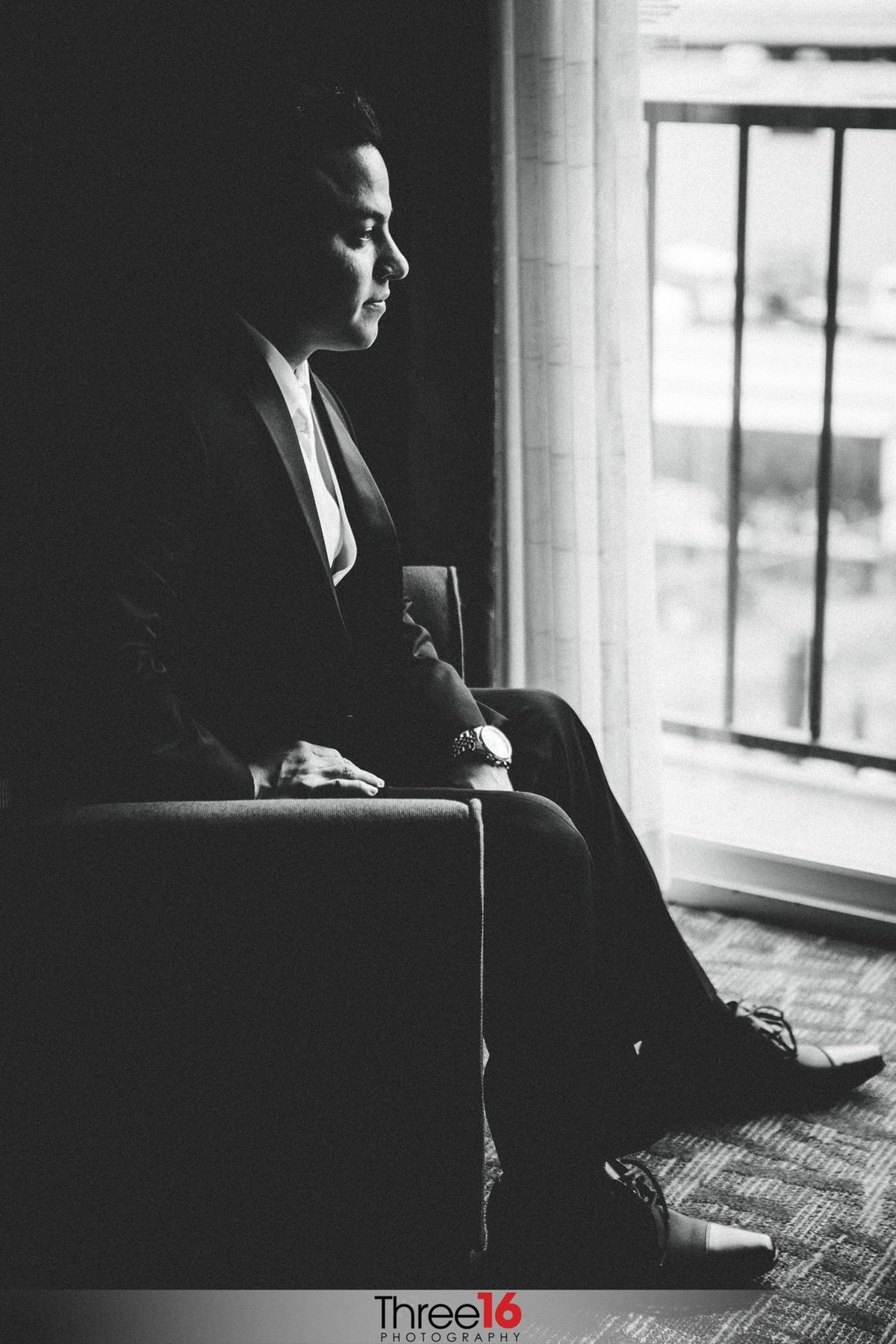Black & White photo of the Groom sitting in a chair looking out the window in deep thought before his wedding ceremony