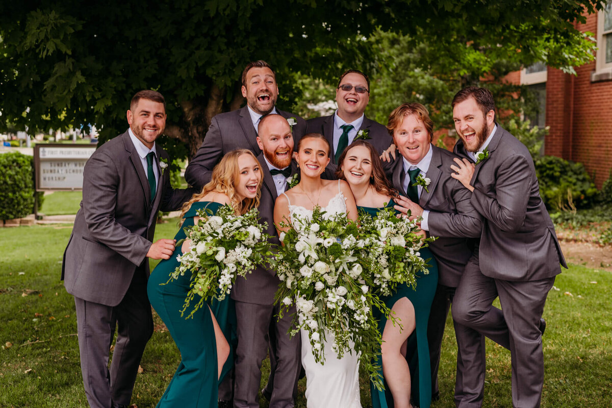 photo of a bride groom and their wedding party in a big group hug while everyone smiles in front of a tree