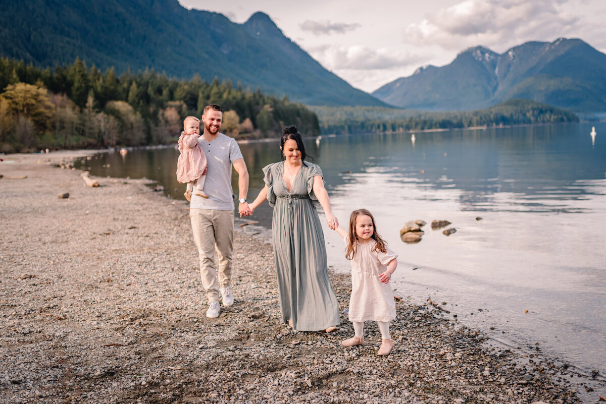 Family in a line holding hands young girl leading them. Family dressed in white, light blue, green and pink. Mountain, beach setting and lake behind them.