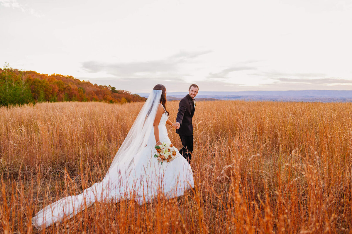 Photo of a groom holding his brides hand and walking into a field at sunset