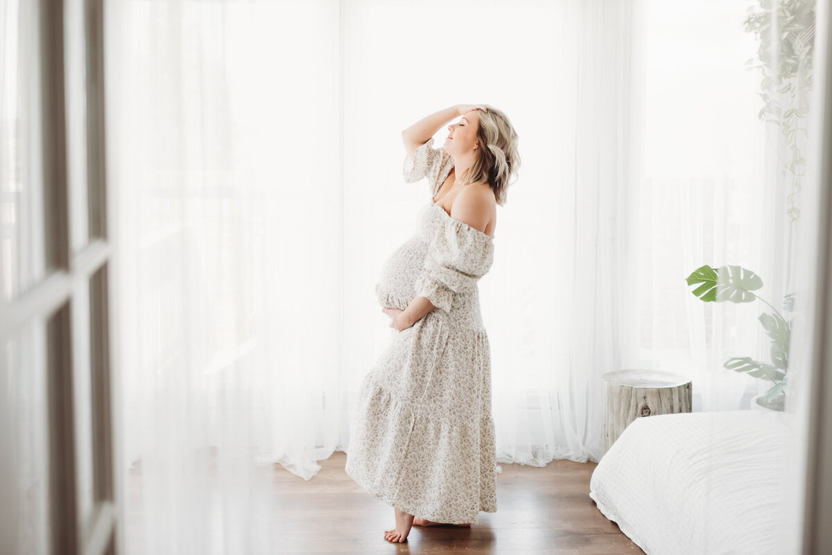 pregnant maternity photographer standing in sunroom with one hand on belly and one hand in hair