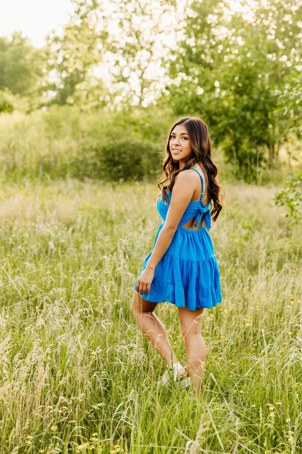 stunning high school senior in a bright blue dress looking back as she walks away in tall grass in a field near Oshkosh