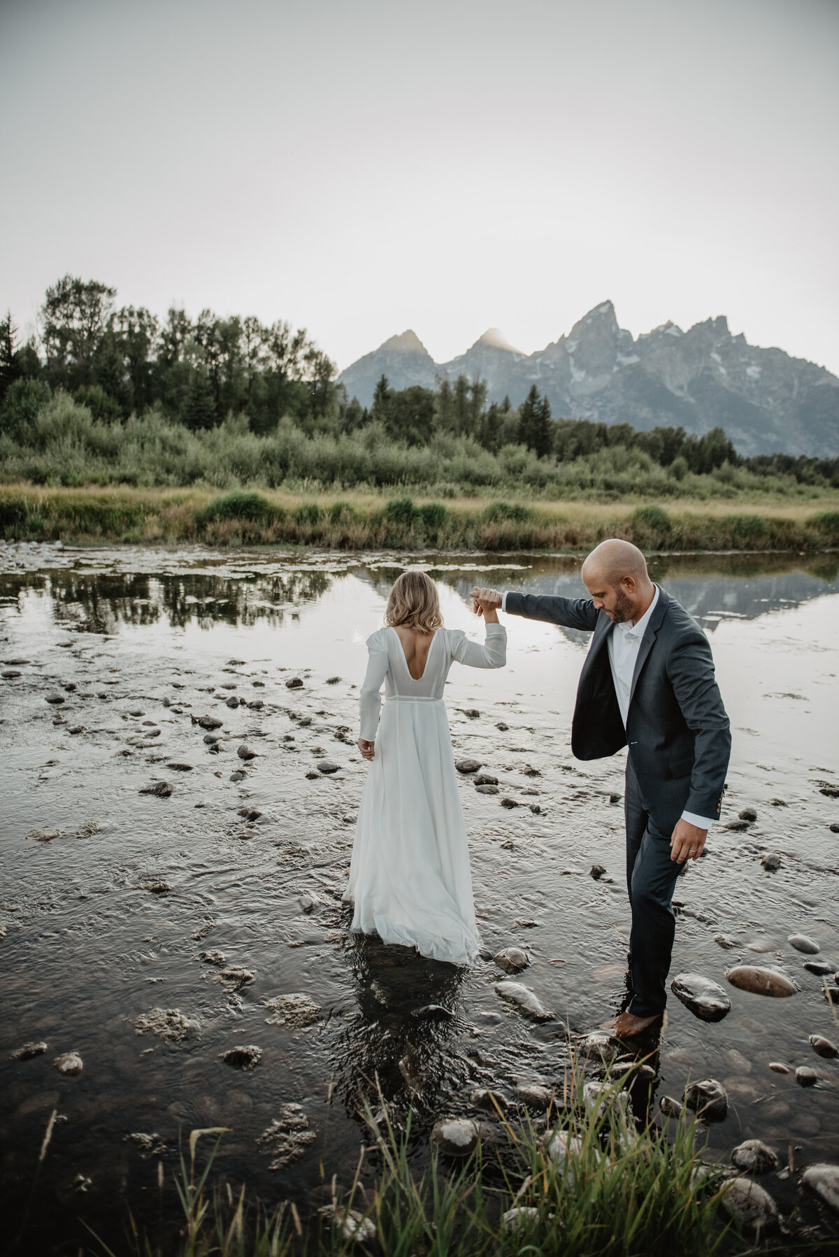 jackson wyoming photographer captures adventure elopement session in the Tetons with the bride and groom dancing in the river among the rocks and brush for their grand teton wedding pictures